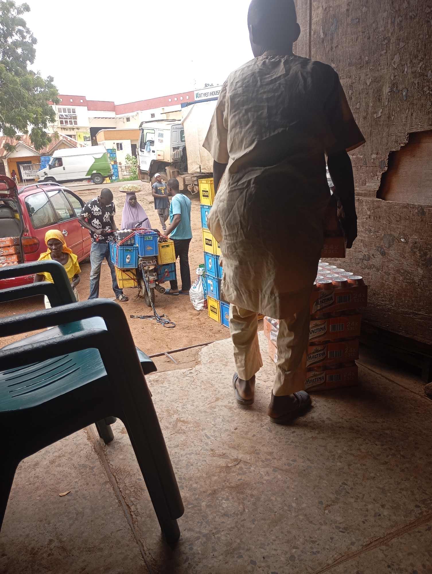 In an outdoor, likely urban environment, a person stands with their back to the camera, wearing a loose-fitting, light-colored tunic and pants. They are standing on a concrete ground, next to stacks of orange and white beverage crates. In the mid-ground, several people are gathered around a bicycle-powered cart laden with blue and yellow crates. A red car is parked on the left. In the background, buildings and a white truck are visible. The scene suggests a marketplace or delivery point.