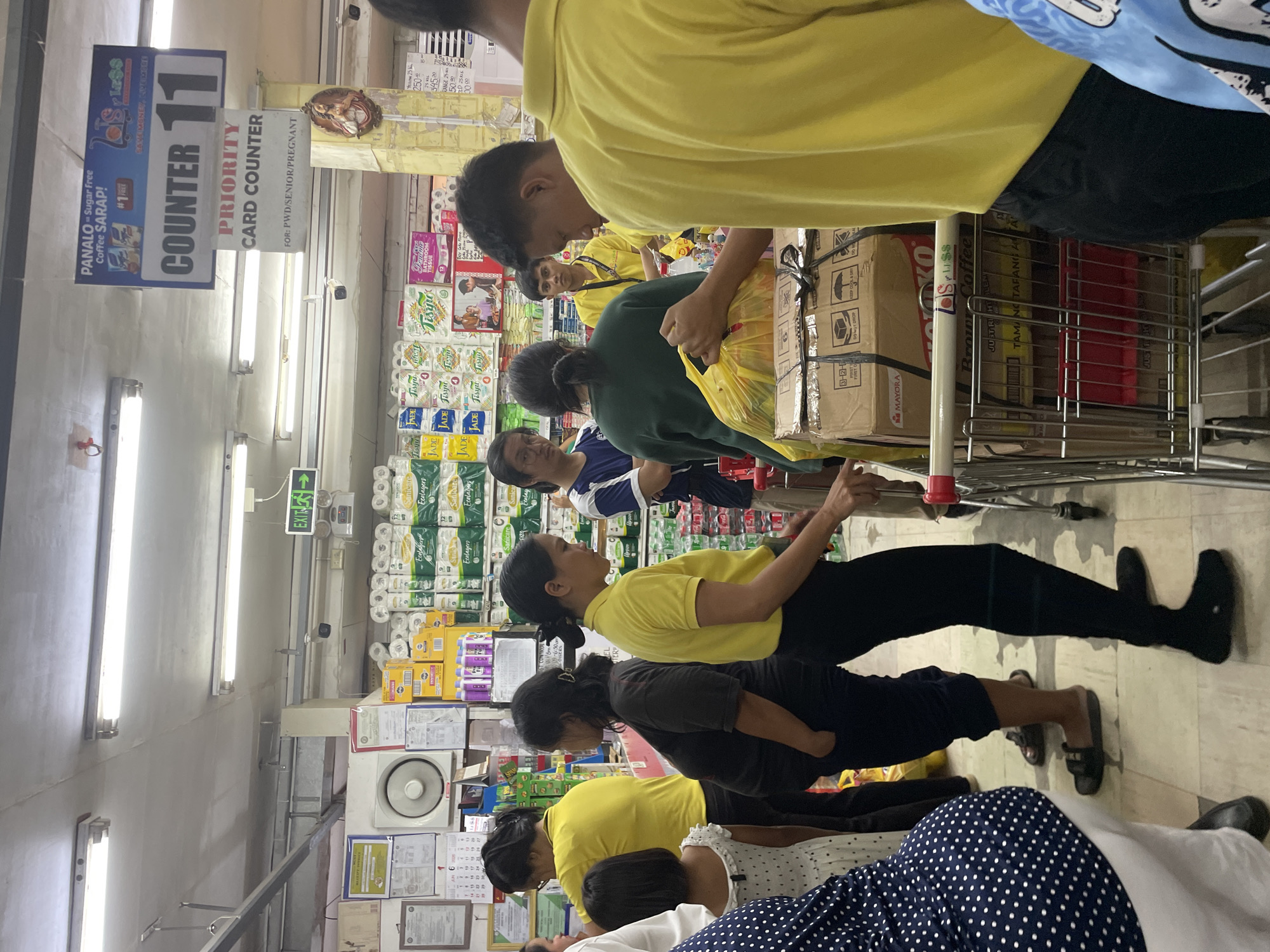 This image captures an active indoor scene at LS Supermarket, a retail establishment located in Davao City, Philippines. The primary focus is a checkout area, specifically "COUNTER 11," which is designated as a "PRIORITY CARD COUNTER FOR: PWD/SENIOR/PREGNANT" as indicated by a prominent overhead sign.
The environment features a light-colored ceiling with exposed metal conduits and multiple fluorescent light fixtures, providing bright illumination. A white wall-mounted fan, several framed documents or certificates, and a calendar displaying "JUN 6" are visible on the wall behind the customers on the left. An "EXIT" sign with a green arrow is positioned on the upper right side of the frame. A CCTV camera is mounted near the exit sign. A religious icon, possibly of the Virgin Mary, is affixed to a yellow column on the right. Shelves stocked with various products, including multiple brands of tissue paper (e.g., Sanicare Ecolayers, Jade, Tesyon) and purple-bottled items, are visible in the background. An advertisement for "PANALO = Sugar Free Coffee SARAP!" bearing the "LS Less" logo hangs near the counter sign.
Approximately ten to twelve individuals are present, primarily adults, along with one child. Many people are wearing yellow shirts, suggesting a possible uniform for employees or a large group. In the foreground left, a person wearing a dark blue polka-dotted top and white lower garment is partially visible, facing away. Adjacent is a small child in a white polka-dotted dress. A woman in a yellow shirt and black pants stands facing the counter, followed by a woman in a dark brown/black shirt and dark pants, who appears pregnant and has her hands in her pockets. Another woman in a dark green long-sleeved shirt stands behind a metal shopping cart filled with cardboard boxes (one visibly branded "MAYORA") and a yellow plastic bag, handling items. To her right, a man in a yellow shirt and dark pants is processing or packing items from a separate shopping cart that displays the "LS Less" logo on its handle. Further to the right, a man in a dark blue shirt is partially visible. In the background, another man in a yellow shirt observes the scene. The individuals are engaged in queuing and transactional activities typical of a supermarket checkout line. The floor is light-colored tile.