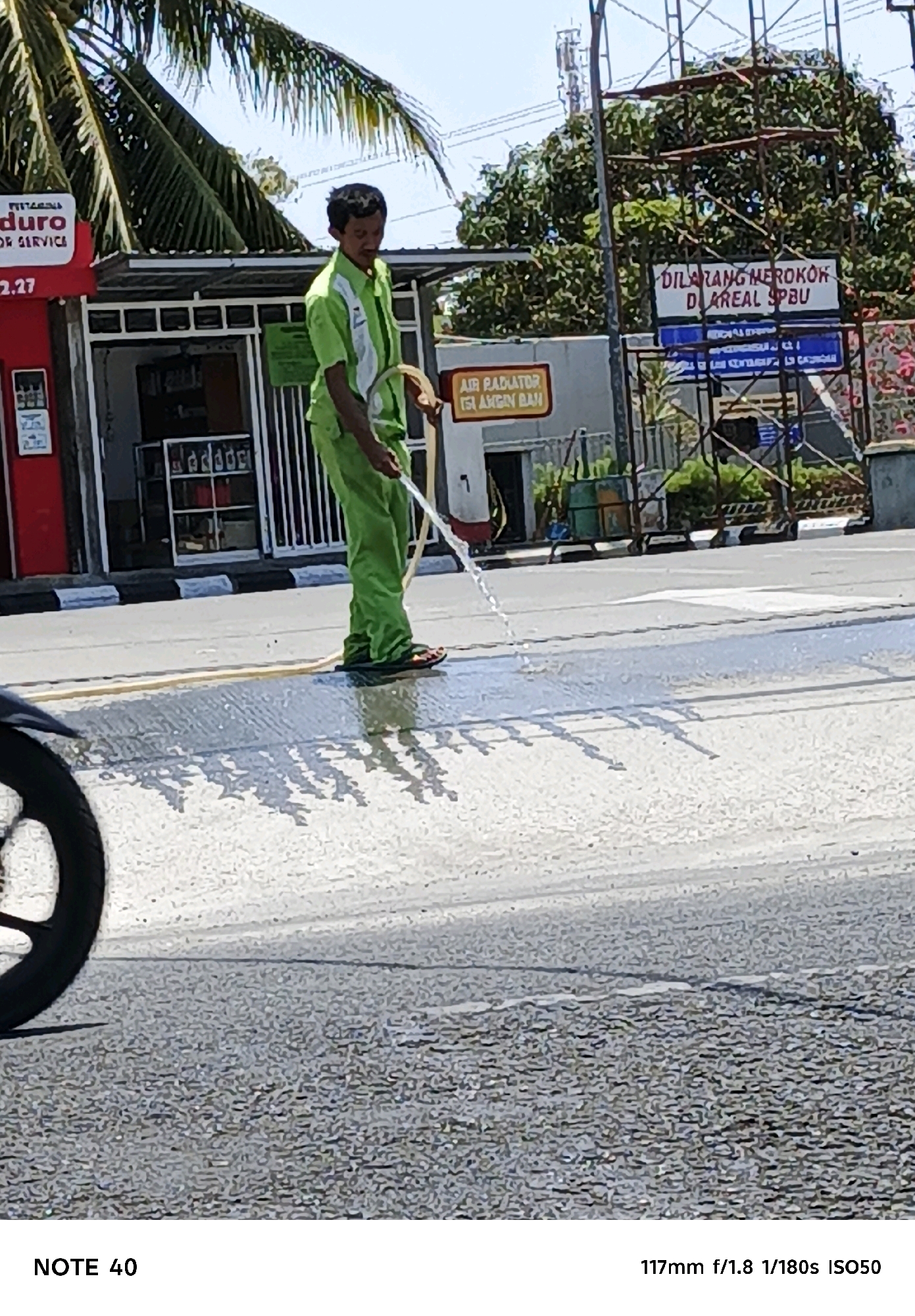In the city of Cilacap, Indonesia, a man wearing a green uniform is watering the street from a hose. He is standing on the road, which appears to be wet and reflecting the sunlight. In the background, there are buildings, trees, and scaffolding. A red sign with the text "DURO" and "FOR SERVICE" is visible on one of the buildings. Another sign in the background reads "DILARANG MEROKOK DI AREA SPBU", which translates to "SMOKING IS PROHIBITED IN THE GAS STATION AREA". A motorcycle's front wheel is partially visible on the left side of the image. The scene suggests a cleaning or maintenance activity taking place outdoors.