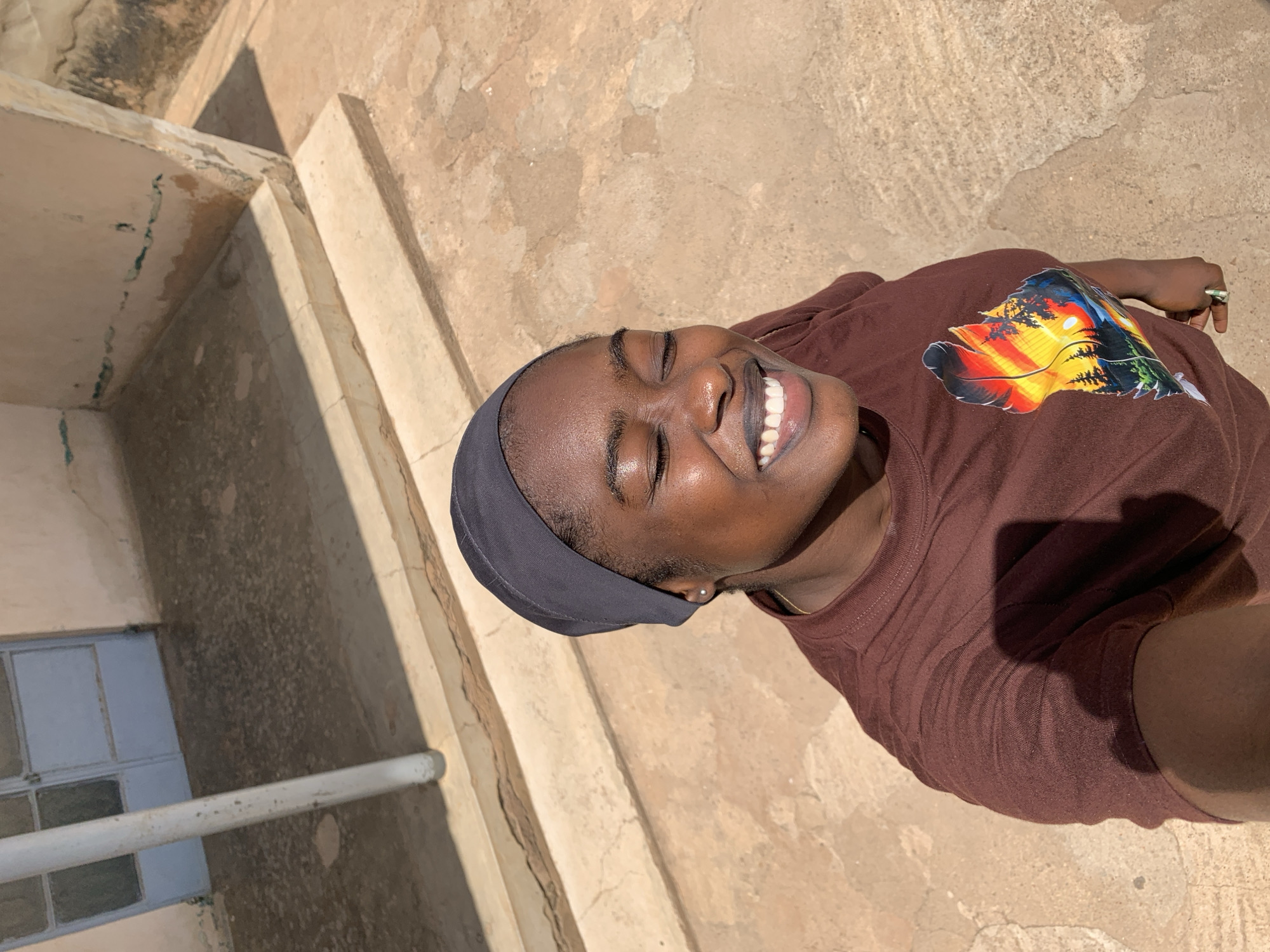 The image depicts a person with dark skin and a black head covering, smiling with closed eyes. They are wearing a brown t-shirt with a colorful design on the right shoulder. The background consists of a textured, light-brown surface with visible cracks and variations in color, possibly concrete or stone. There are elements of a building structure visible on the left, including what appears to be a window. The location is the New Stadium, Nigeria.
