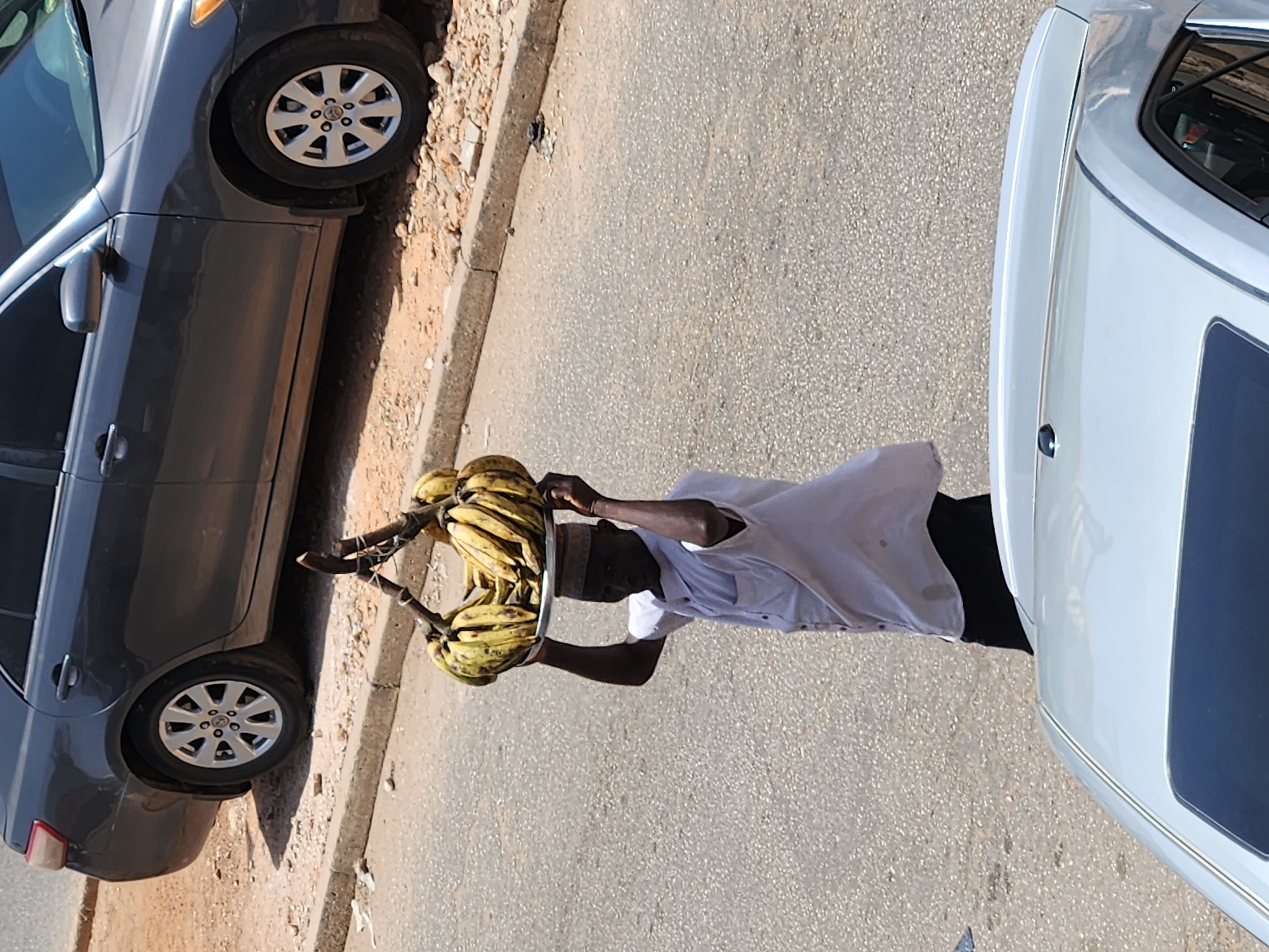 A man is walking on a road in an unidentified city with bunches of bananas on a tray atop his head. The man is wearing a white t-shirt and black pants. To his right is a dark gray sedan parked on the side of the road. To his left is a white car parked on the side of the road. The road appears to be asphalt. The edge of the road where the dark gray sedan is parked appears to be cracked with patches of dirt. The weather seems to be sunny.