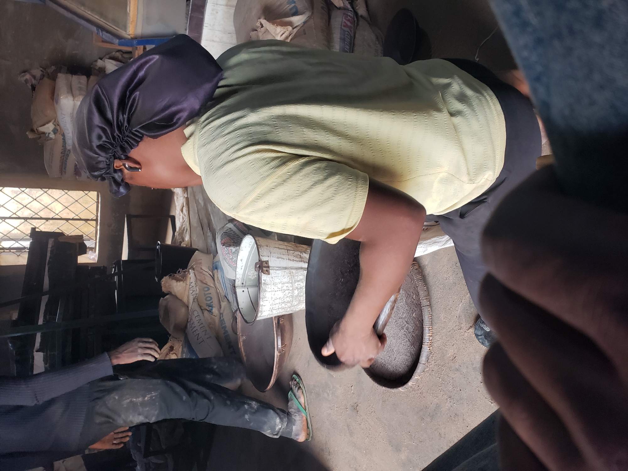 The image depicts a scene inside what appears to be a workshop or storage area, likely in Jos, Nigeria. In the foreground, a person is kneeling and holding a dark, shallow pan, seemingly sifting or processing something on a woven mat. This person is wearing a yellow, textured shirt and a dark satin bonnet. To the left, another person stands wearing dark pants with white smudges and a dark long-sleeved shirt, also with smudges on their pants. Their footwear is a green flip-flop.
The environment is cluttered with various objects. Several large sacks, some with visible branding that could indicate cement or similar materials, are stacked in the background. A distressed white metal bucket is visible between the two individuals. There are large, dark, circular pans on the floor. A window with diamond-shaped panes is present in the background, allowing some light into the space. The floor appears to be concrete. The overall impression is of a functional, possibly industrial or craft-related space, given the materials and tools present. The presence of cement bags and the sifting action suggest a process related to construction materials or a similar trade.
