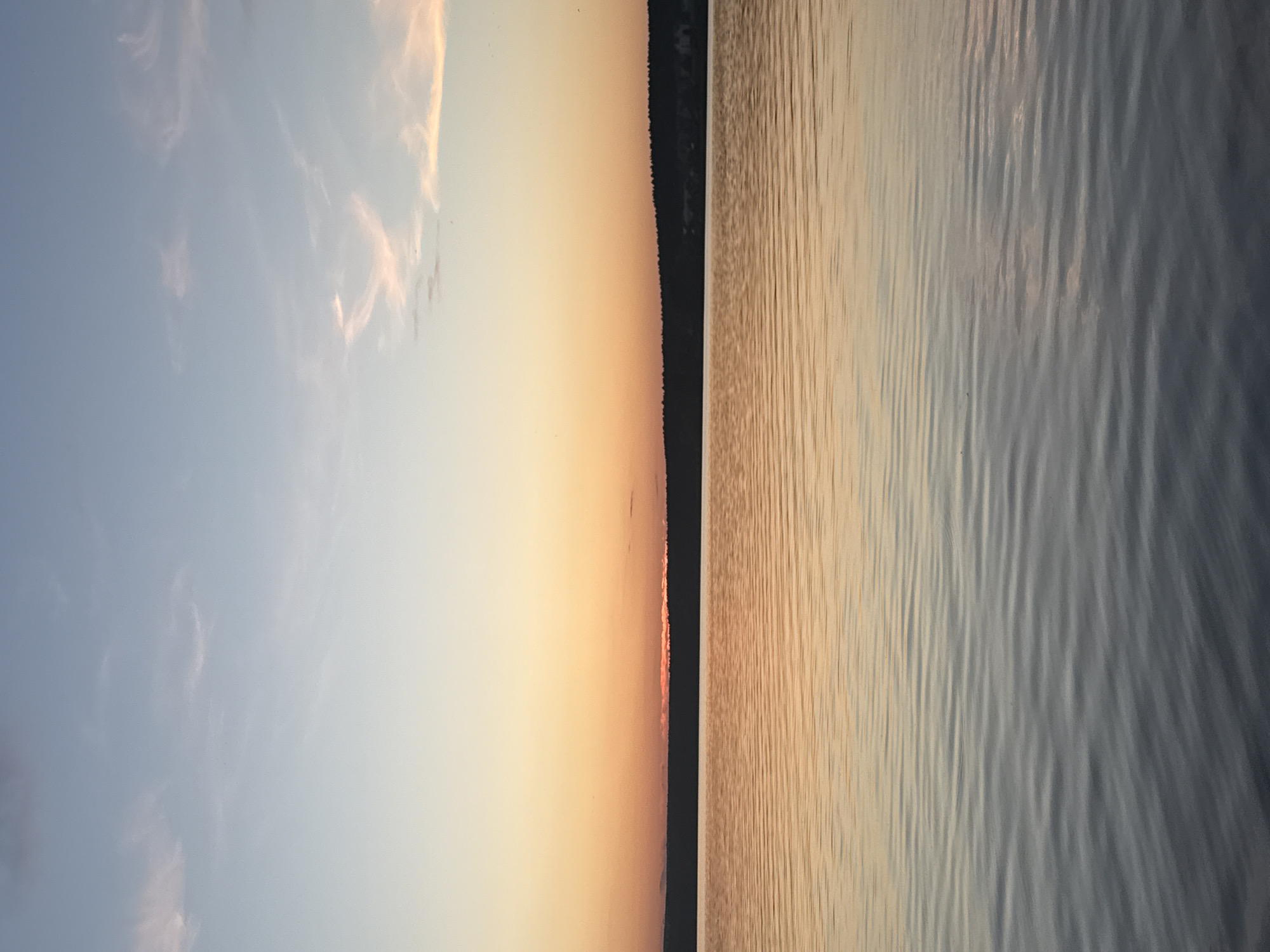 The image depicts a serene sunset over a large body of water, likely the Votkinsk Reservoir in Votkinsk, Russia. The upper sky displays a pale blue, transitioning horizontally into a vibrant gradient of light yellow and orange-red closer to the horizon. Sparse, elongated cirrus clouds, faintly tinged with pink, are visible across the lighter blue and yellow sections of the sky.
A distinct, dark horizontal silhouette of a distant landmass, consistent with a densely forested bank, defines the horizon line. Directly above the central portion of this landmass, a concentrated band of intense orange-red light indicates the position of the setting sun, partially obscured by the distant terrain. Subtle, lighter-toned areas are discernible within the silhouette on the far right, suggesting potential distant structures or clearings.
The lower half of the image is occupied by the water, which reflects the sky's colors. The water surface exhibits numerous small, parallel ripples, indicating a calm but not entirely still condition. The coloration of the water transitions from a warmer orange-yellow nearest the reflected horizon to a cooler, desaturated blue-grey in the foreground. No human figures, animals, or man-made objects such as boats or structures are present in the immediate or mid-ground of the water or the visible shoreline. The scene is devoid of any explicit actions or interactions.