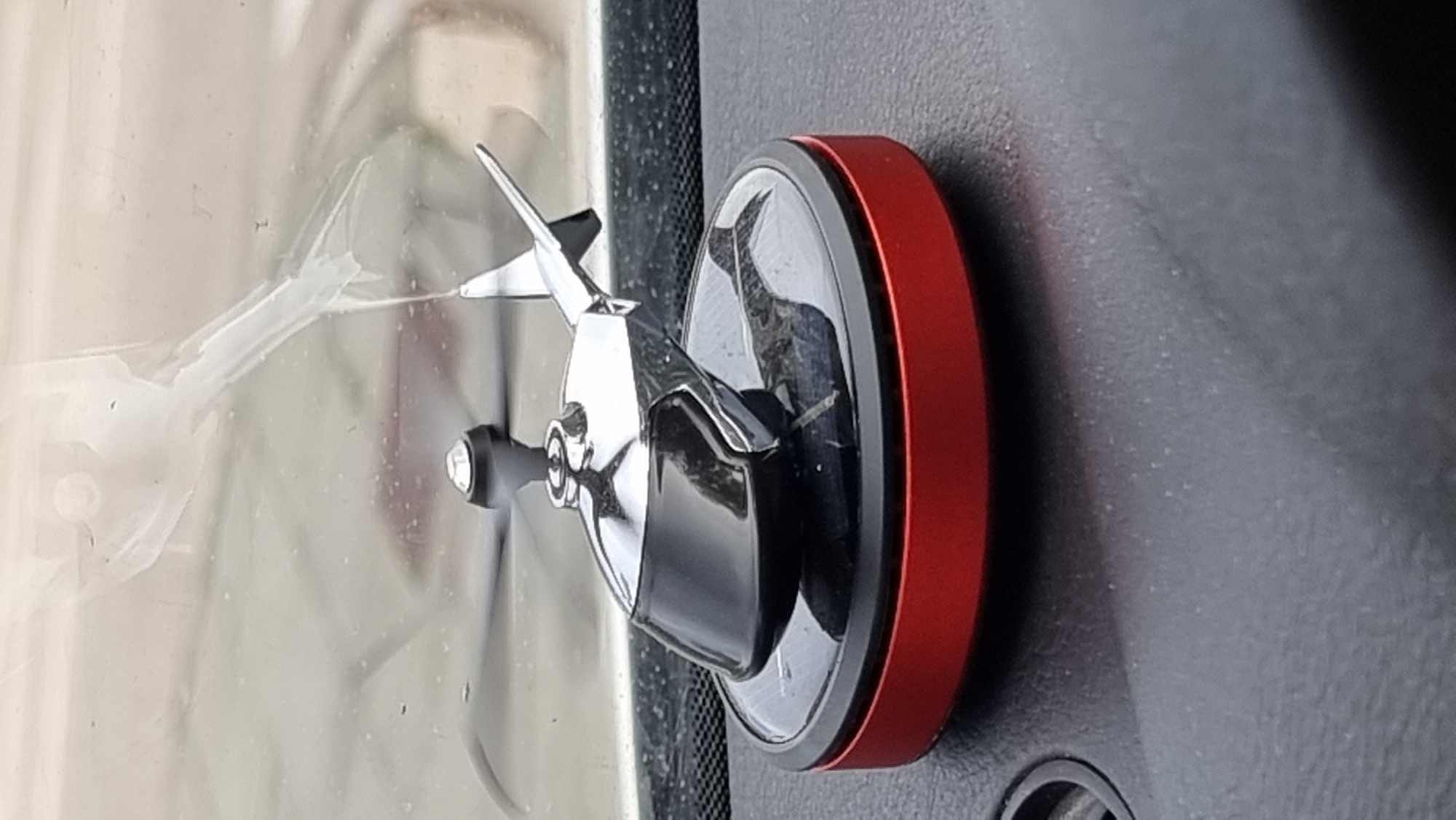 A chrome and black helicopter-shaped car air freshener with spinning blades sits on a red and black base. The base appears to be solar-powered, with a solar panel visible. The air freshener is positioned on a dark, textured surface, likely the dashboard of a vehicle. The background is out of focus, showing a blurred outdoor scene with a greyish-brown hue and some light-colored streaks, possibly reflections or dirt on a window. There is a faint, white, elongated shape in the upper portion of the image, which could be a reflection or a sticker on the window. The scene is captured from a slightly elevated angle, looking down at the air freshener. The image does not depict any people or specific identifiable landmarks of Sant Joan les Fonts, Spain.