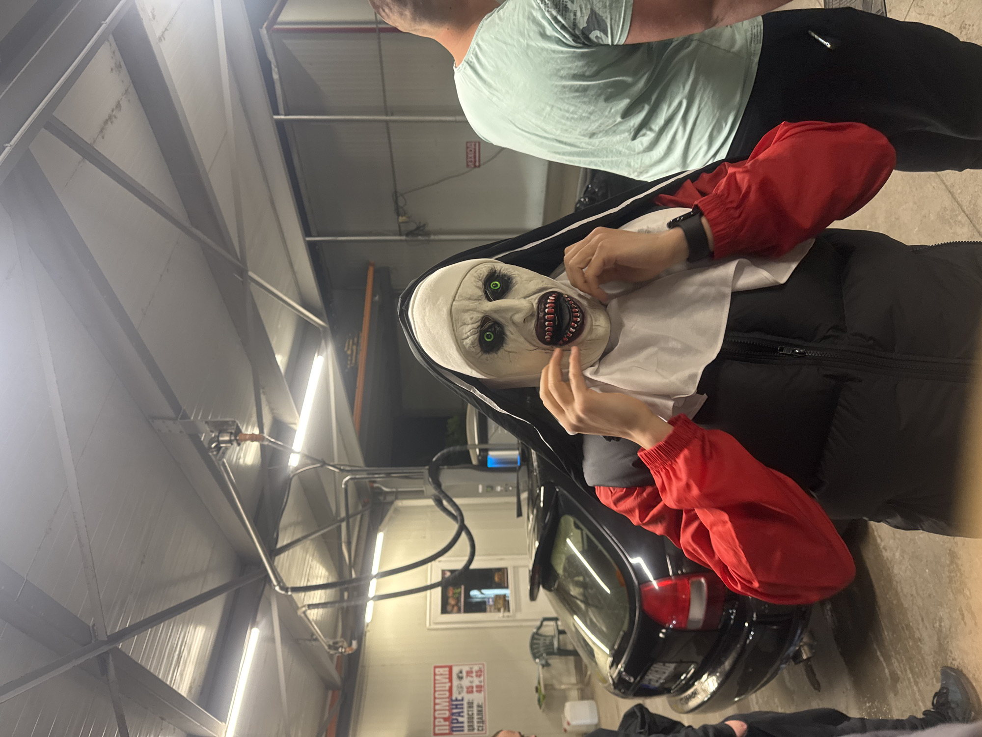 The image depicts an indoor self-service car wash bay, featuring a person in a terrifying "Nun" costume, reminiscent of the character Valak from "The Conjuring" films. The white mask has striking green eyes, dark features, and exposed, sharp teeth. The person wears a black nun's habit, a black puffer vest, and bright red long sleeves or gloves, with their hands positioned near the mask.
Above and behind the costumed figure, another person is partially visible from the back, wearing a light blue short-sleeved shirt and dark pants. The legs and feet of a third individual, dressed in dark clothing and black sneakers, are seen at the bottom of the frame.
A black car, with its rear section and taillights visible, is parked within the bay. The environment is industrial, characterized by a concrete floor, metal beams, corrugated panels on the ceiling and walls, and bright fluorescent lighting. Car wash hoses and equipment are also present. A red sign on the wall displays Cyrillic text: "ПРОМОЦИЯ ПРАНЕ" (PROMOTION WASHING), followed by pricing details in Bulgarian Leva (e.g., "ЦЕННОСТИ: 80 - 70 - 60 лв.", "СЕЛЕСТИЯ: 40 - 45 - 30 лв."). Another partially visible sign reads "ИЗХОД" (EXIT). The scene, likely taking place in Plovdiv, Bulgaria, suggests a playful or themed event at the car wash.