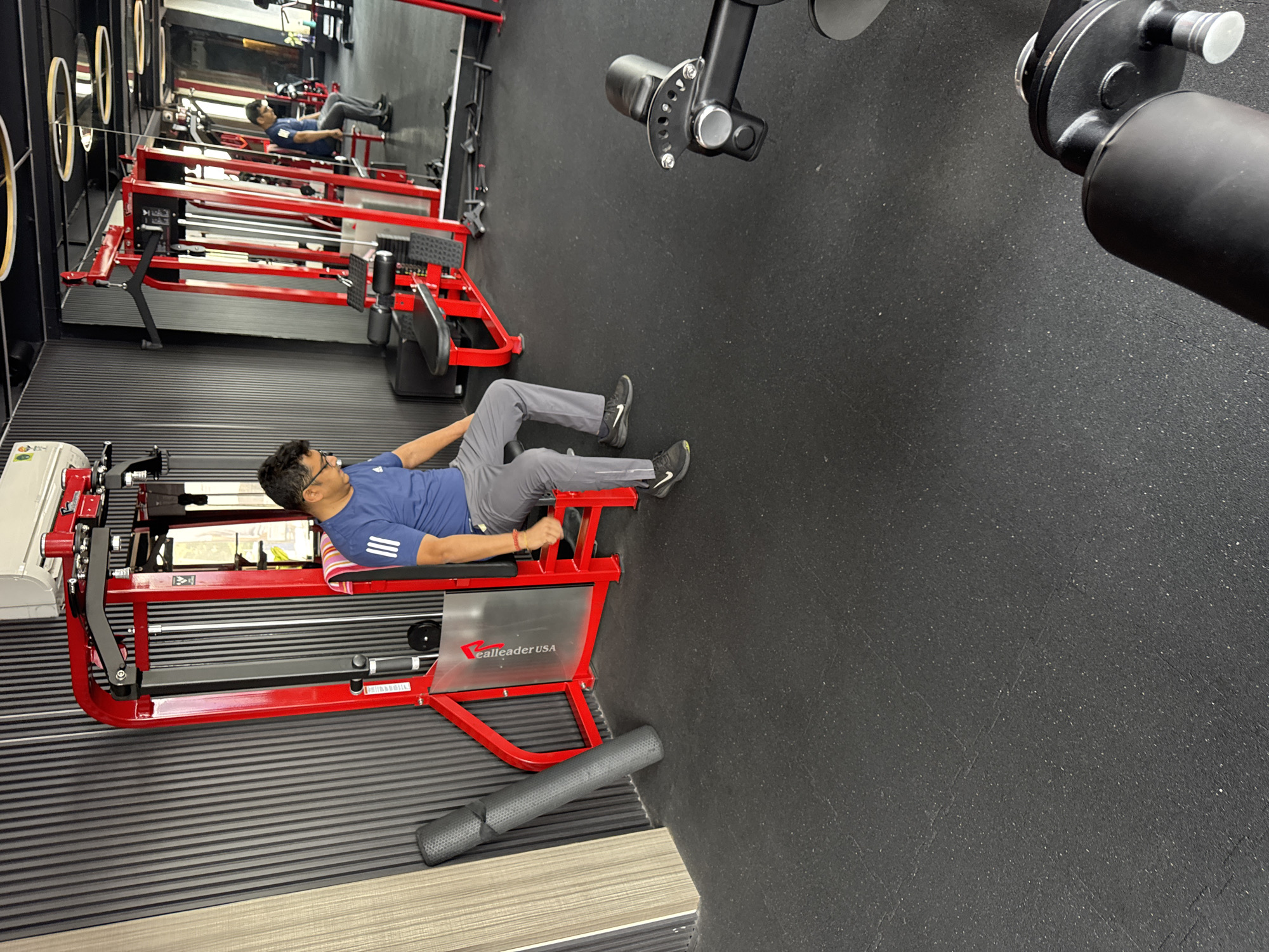 The image captures a scene in a gym in Kolkata, India. A man wearing glasses, a blue t-shirt, and gray pants is positioned on a red and gray weight machine. He is lying on his back with his legs bent at the knees and his feet placed on the machine's footrests. The machine is labeled "ealleader USA". The gym has black flooring and mirrored walls with decorative circular elements. In the background, other weight machines are visible.