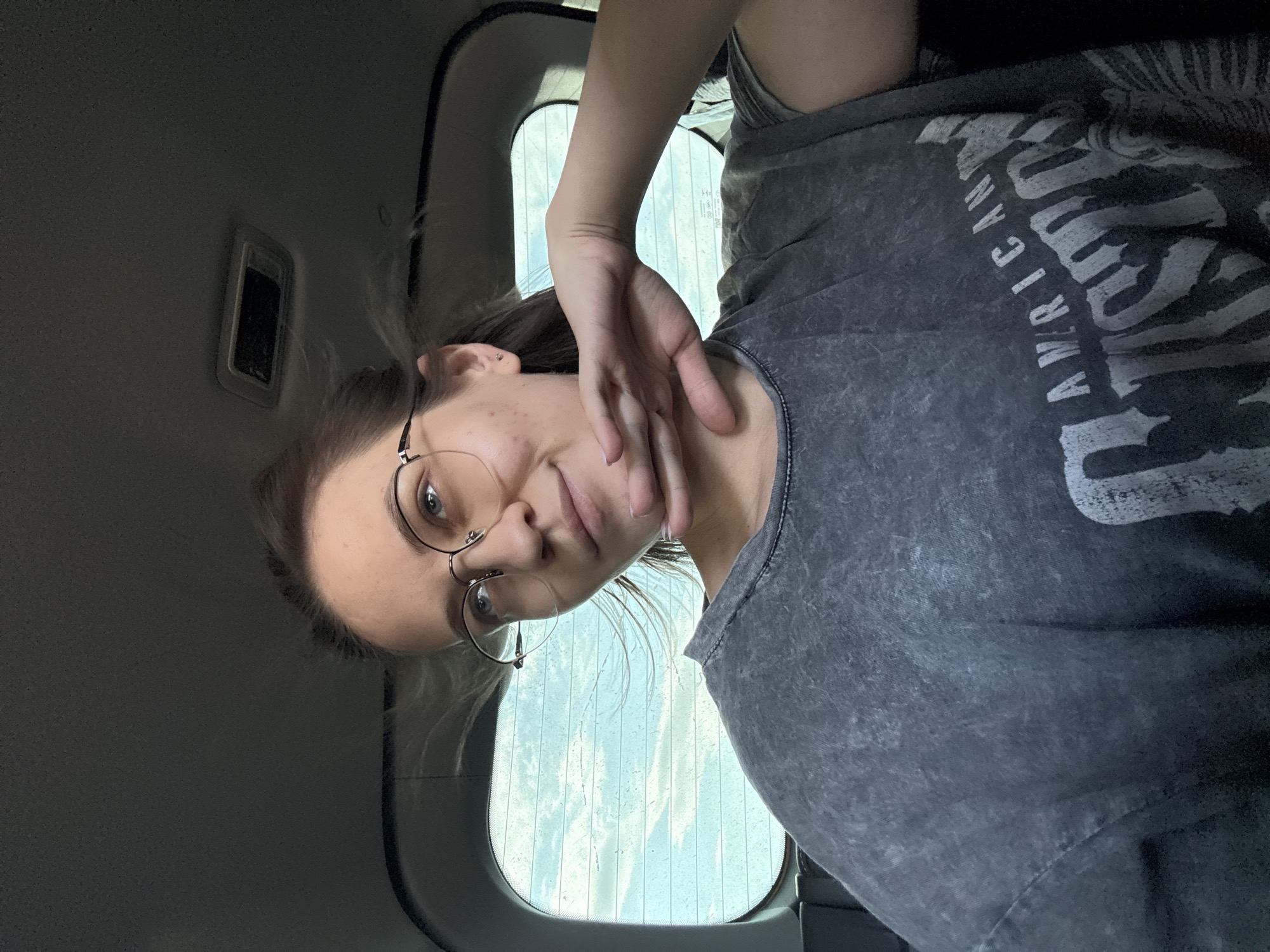 The image shows a person inside a vehicle, likely a car. The person has light skin, light hair pulled back, and wears round glasses. They are wearing a gray t-shirt with white lettering, part of which is visible and appears to spell "AMERICAN". The person's hand is near their face. The car interior is dark, with a window behind the person showing a blurry exterior. There is a vent visible in the car's ceiling. The location is not identifiable as being in Berezka, Russia, as there are no distinguishing features visible in the image that would indicate a specific geographic location.