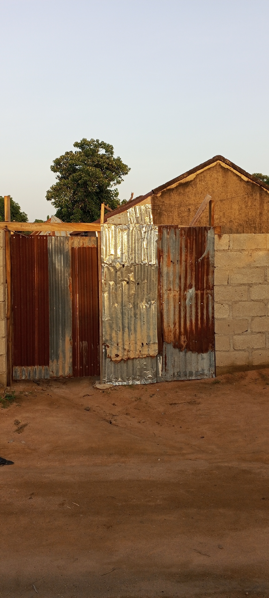 The image shows a corrugated metal gate and a section of a building in Jalingo, Nigeria. The gate is made of several sheets of corrugated metal, some of which are rusty and discolored. To the right of the gate is a wall constructed from cinder blocks. Behind the gate and to the right is a building with a sloped, tiled roof and plastered walls. A large tree with green foliage stands behind the gate and to the left of the building. The ground in front of the gate is made of reddish-brown earth, with some scattered debris and small plants. The sky above is a clear, pale blue, indicating daytime. The scene appears to be an exterior view of a residential or commercial property.