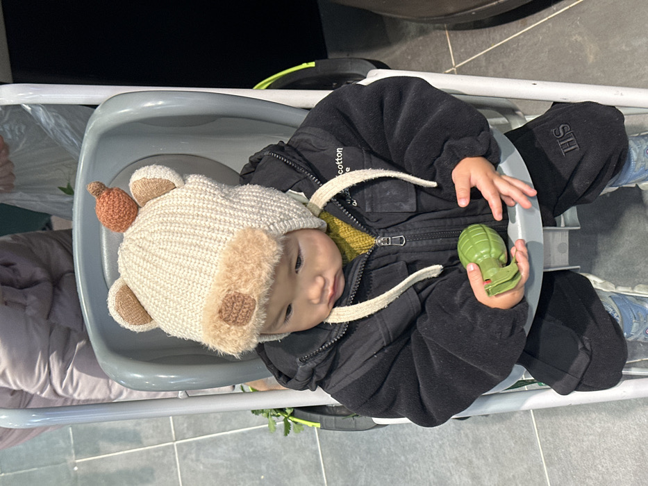 An infant is seated upright in a grey plastic high chair or stroller within an indoor setting. The infant wears a cream-colored knitted hat adorned with bear ears, a brown faux fur band, and a small orange knitted carrot detail on the top, with ties hanging under the chin. A dark, likely black, zip-up jacket is worn over a yellow knitted sweater. The infant's black pants feature the embroidered initials "HS" on the left leg, and light blue sneakers with white soles are visible. The infant holds a bright green plastic toy grenade in the right hand, while the left hand rests on the high chair's front tray. The infant's gaze is directed downward and to the left. In the upper left background, a portion of an adult figure wearing a light-colored padded jacket is visible. A green leafy object is partially seen to the left of the high chair, and a translucent plastic bag is present in the upper right background. The floor is tiled with dark grey square tiles. The specific location within Jiangjin, China, is not discernible from the image.