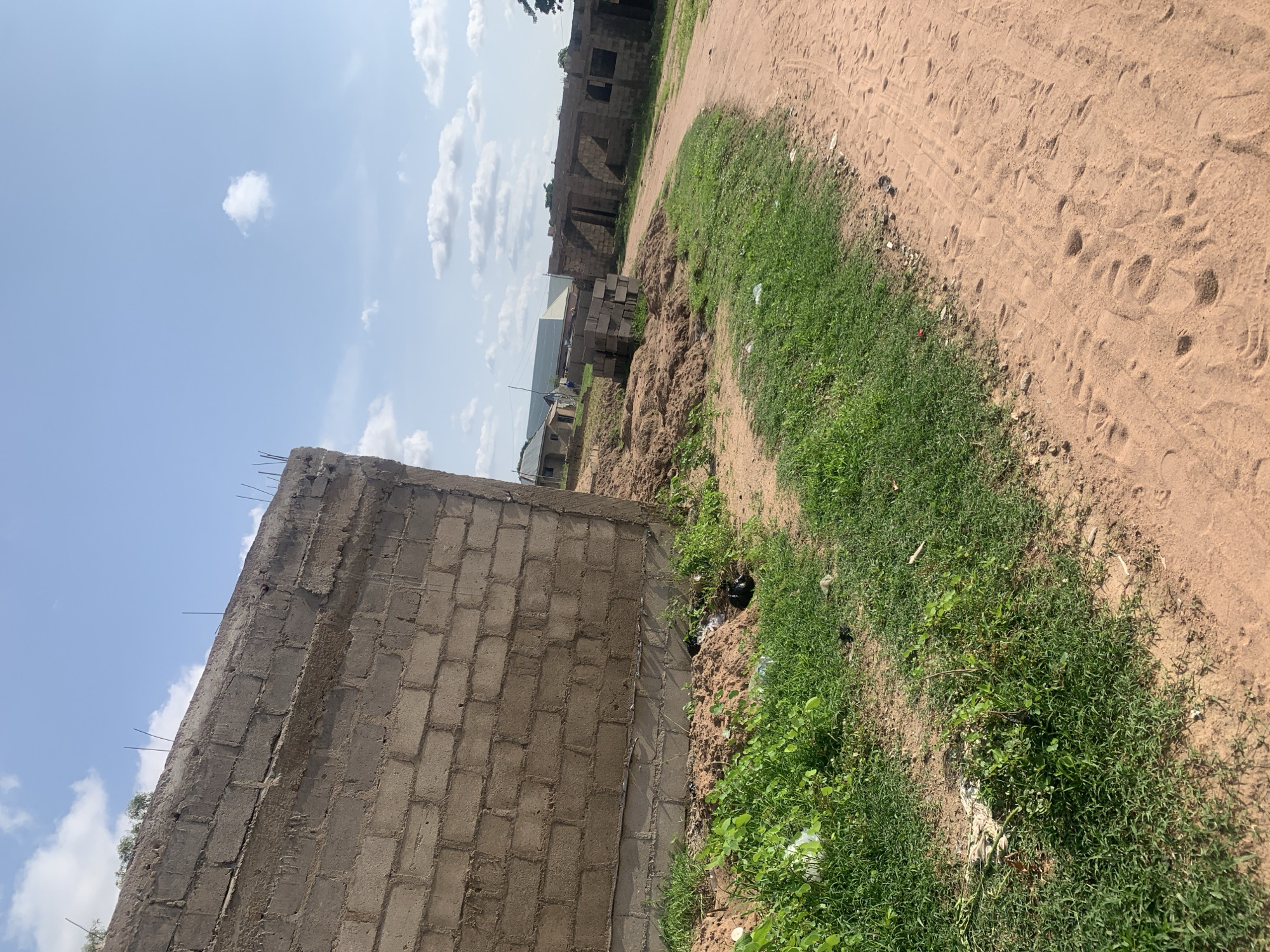 An eye-level, medium shot captures a dirt road and a construction site under a partly cloudy sky in Jalingo, Nigeria. The foreground is dominated by a dirt path, marked with footprints and tire tracks, leading into the distance. To the left, a patch of green grass grows beside the road, with a partially constructed brick wall rising prominently. The wall is made of concrete blocks, with exposed rebar protruding from its top edge. Further back, the construction site reveals more unfinished brick buildings, some with open doorways and windows. Piles of dirt and scattered building materials are visible among the structures. Distant homes with roofs are also visible, suggesting the site is in a developing residential area.