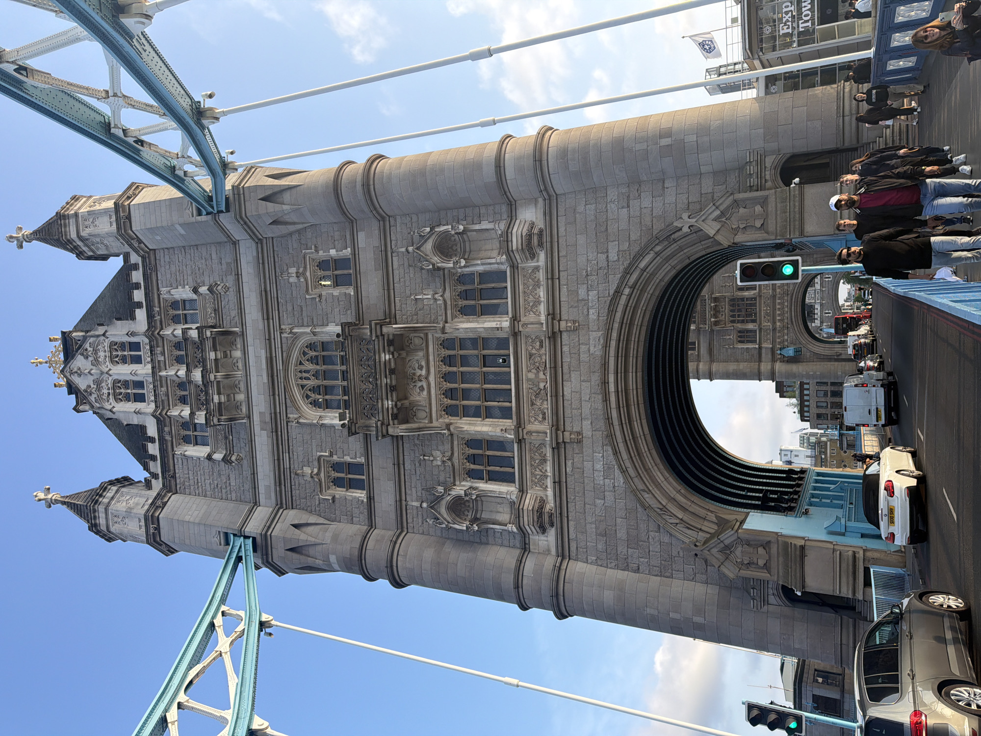 The image shows the Tower Bridge in London, UK. The bridge's architectural details, including its towers, windows, and archways, are visible. Several people are walking on the bridge, and cars are driving underneath. The sky is partly cloudy. The traffic light is showing green.