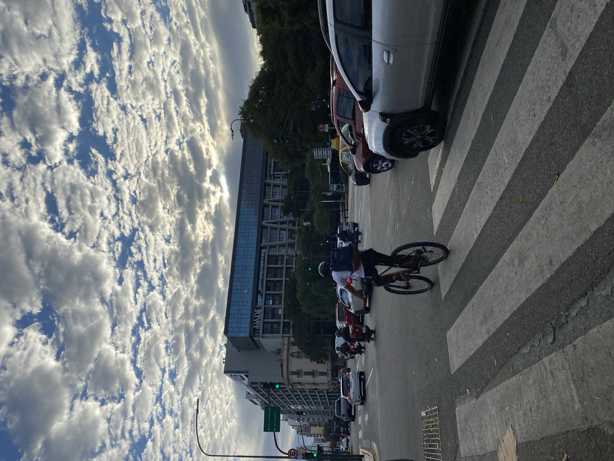 The image depicts a street scene in Buenos Aires, Argentina. In the foreground, a cyclist crosses a zebra crossing. A line of parked cars is visible on the right side of the street. In the background, a modern building with the letters "UADE" is seen, along with other buildings and a street sign indicating "Av. Pte. Peron" and "Avellaneda". The sky is filled with clouds.
