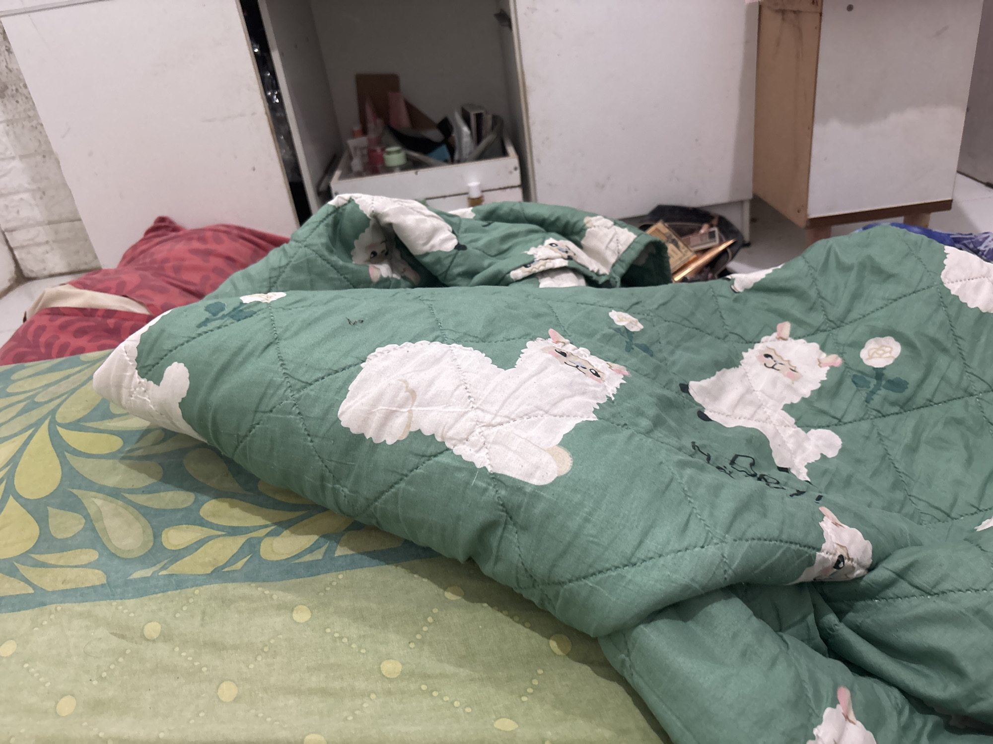 The image depicts a close-up view of a bed with a green comforter featuring a pattern of white sheep. The comforter is partially folded, revealing a light green sheet with a leaf-like pattern underneath. In the background, to the left, is a white wardrobe or cabinet, and to the right, part of a light brown wooden cabinet is visible. Various items are stored on shelves within the wardrobe, including bottles and boxes. The scene appears to be indoors, possibly a bedroom, within a residential setting in North Jakarta, Indonesia. There are no people or animals visible in the image.