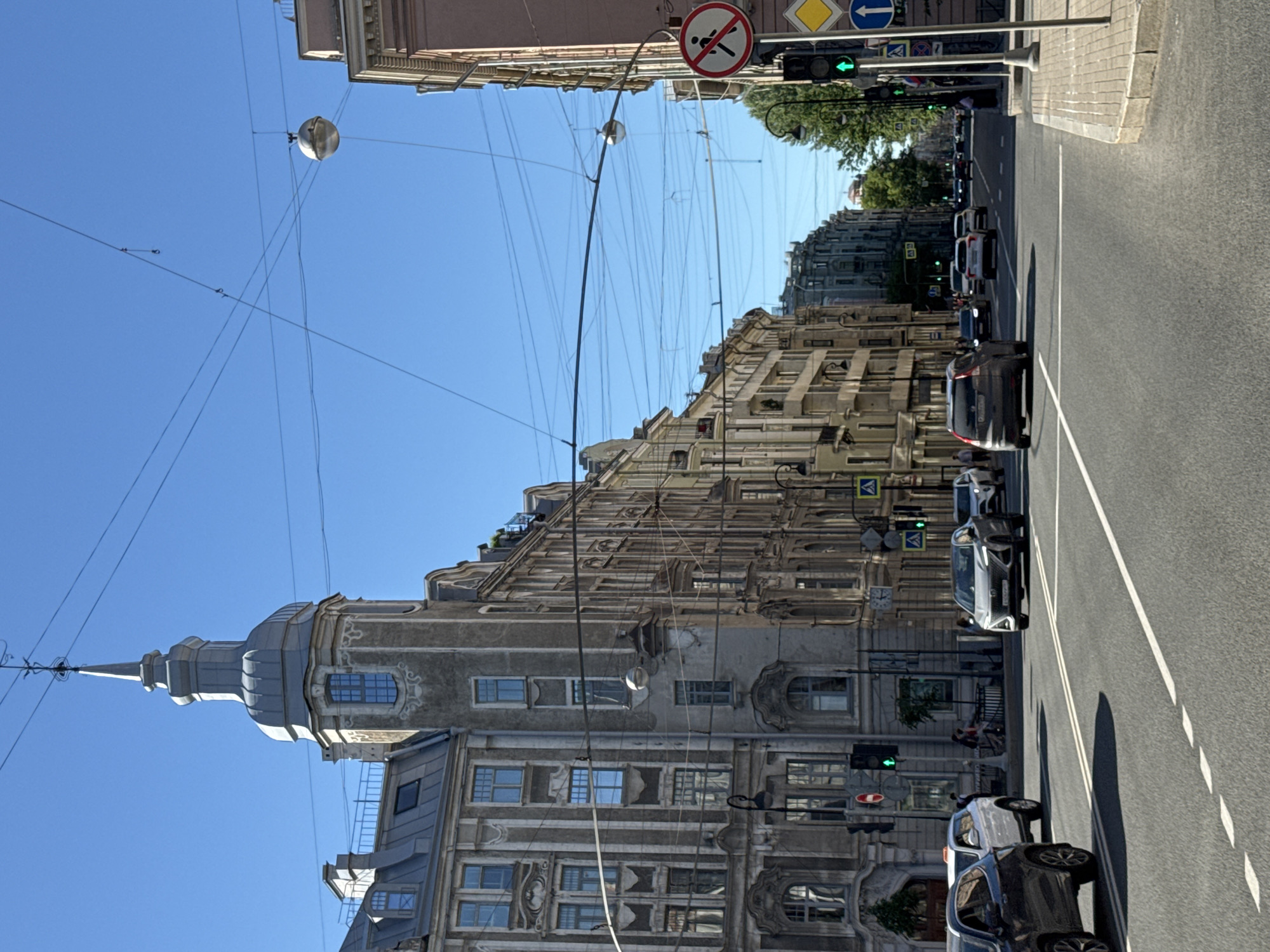 This image captures a wide-angle view of Liteyny Prospekt in Saint Petersburg, Russia, looking approximately south-east from the vicinity of its intersection with Ulitsa Pestelya. The upper third of the image is dominated by a clear, bright blue sky, crisscrossed by numerous thin black overhead wires, likely for electric public transport (trolleybuses/trams) and other utilities. Several spherical, grey streetlights are suspended from these wires. On the left side of the frame, a multi-story building with a distinctive corner tower featuring a grey, metal-clad dome and a slender spire is prominently visible. This building is identified as 29 Liteyny Prospekt. Its facade is a muted grey-beige, detailed with pilasters, ornate window surrounds, and decorative stucco patterns. Adjacent to and beyond this building, a continuous line of similar multi-story structures, exhibiting classical and Art Nouveau architectural elements, recedes into the distance along the street. A smaller, spherical streetlight is suspended above the street near the left sidewalk. A traffic light showing a green signal is visible on the left, below which is a red circular "No Entry" sign. The right side of the frame features a multi-story building with a lighter, possibly pale pink or beige facade, also exhibiting decorative details. Further down the street on the right, a line of mature green trees borders the road, suggesting a boulevard. On the right sidewalk, a tall metal pole supports multiple traffic signs: a circular "No Pedestrians" sign (white background, red border, black pedestrian figure with a red diagonal line), a yellow diamond-shaped "Main Road" sign, and a blue circular "Ahead Only" sign (white upward-pointing arrow). Below these, a traffic light also displays a green signal. The center of the image shows a wide asphalt road with at least three visible lanes of traffic in each direction. White solid double lines mark the center, and dashed white lines delineate lanes. Several passenger vehicles are present: * In the immediate foreground, a dark-colored SUV is partially visible on the left. * Behind it, a white SUV is discernible. * In the middle distance, a silver/grey SUV faces the viewer, positioned in a left lane. * A white sedan, a dark sedan, and other indistinguishable vehicles are further down the road, all appearing to be modern models. Vehicles are either stopped or moving slowly, consistent with urban traffic. Shadows cast by vehicles are visible on the road surface, indicating bright sunlight. A few indistinct human