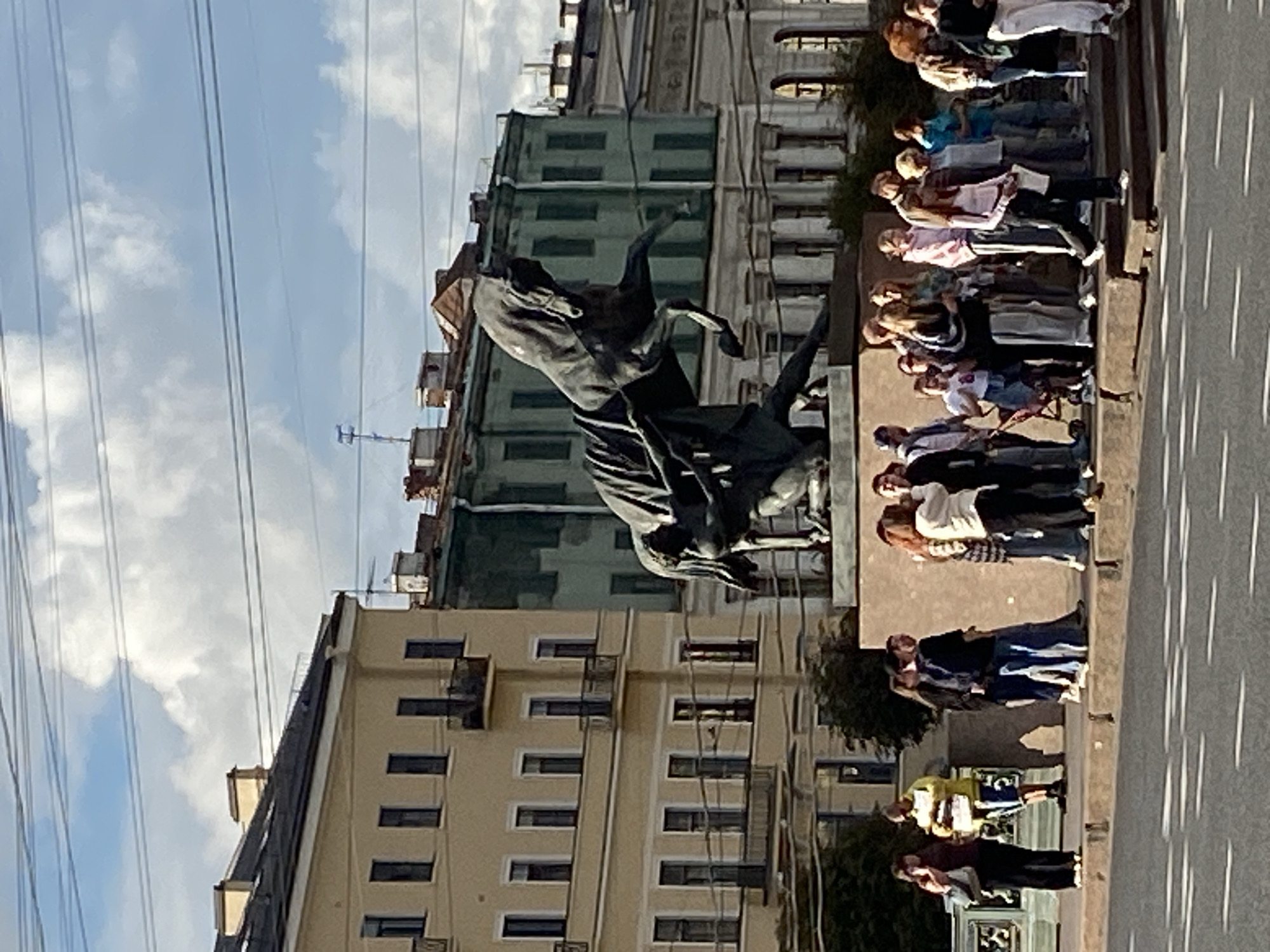 The image depicts a bronze equestrian statue of a man wrestling a horse, commonly known as "The Bronze Horseman," located in Senate Square in Saint Petersburg, Russia. The statue is situated on a large granite pedestal. In the foreground, a group of people are standing and walking along a sidewalk, with a street and road markings visible. Behind the statue, there are classical buildings with multiple stories, painted in light yellow and green hues. The sky above is partly cloudy, with visible power lines crossing the frame. The overall scene suggests a sunny day with moderate pedestrian activity around the monument.