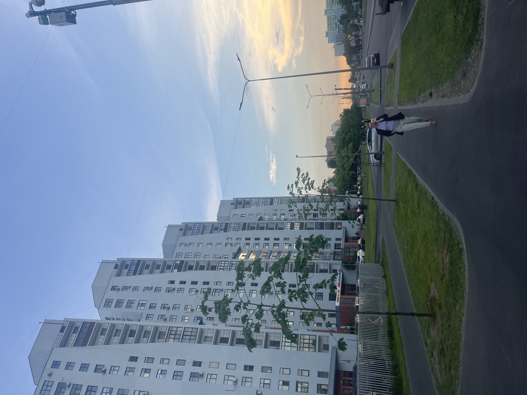 The image captures a street scene in Moscow, Russia, with a focus on modern high-rise residential buildings. In the foreground, a paved pedestrian walkway runs alongside a grassy median. A woman is walking on the walkway, talking on her phone, and wearing a dark blazer over a light-colored top and pants, with sandals. She is positioned to the right of the frame. To the left, a line of tall, light-colored apartment buildings with numerous windows and balconies dominates the background. The sky is a mix of blue and soft orange hues, suggesting it is either early morning or late evening. Streetlights, traffic lights, and utility poles are visible along the adjacent road, where several cars are driving and parked. Trees line the walkway and the roadside, adding greenery to the urban landscape. The overall impression is of a typical urban environment in a residential district during twilight.