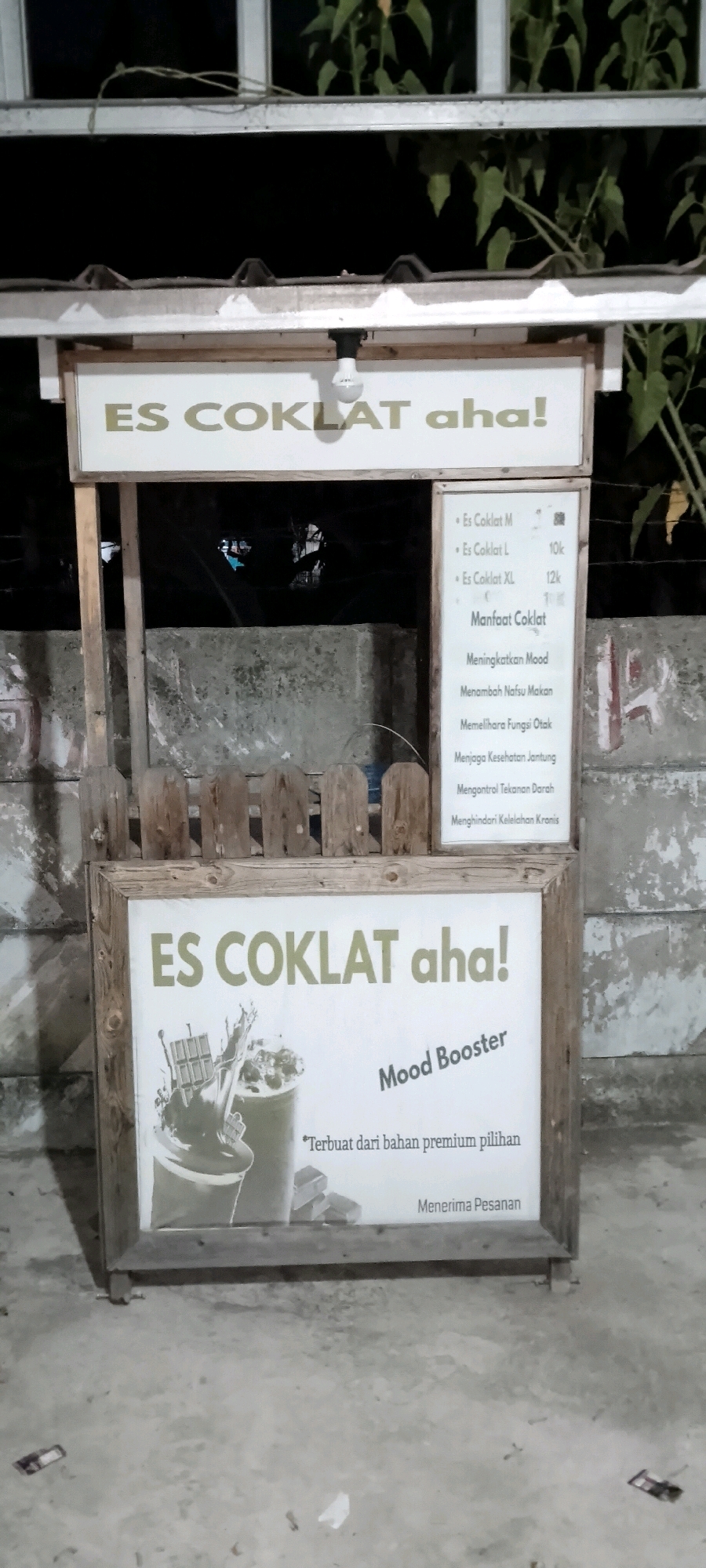 An outdoor, eye-level, medium shot shows a small, wooden food stall at night, likely in the city of Satriamekar, Indonesia. The stall is illuminated by a single bare light bulb hanging from the roof. The stall features two signs. The upper sign, a light-colored rectangle with dark text, reads "ES COKLAT aha!". Below this, a larger sign with a wooden frame displays a large graphic of a chocolate bar and a drink, alongside text. The text on the right side of this larger sign lists sizes and prices for "Es Coklat" (Chocolate Ice), with options for M, L, and XL. It also lists "Manfaat Coklat" (Benefits of Chocolate), including "Meningkatkan Mood" (Boosts Mood), "Menambah Nafsu Makan" (Increases Appetite), "Memelihara Fungsi Otak" (Maintains Brain Function), "Menjaga Kesehatan Jantung" (Maintains Heart Health), "Mengontrol Tekanan Darah" (Controls Blood Pressure), and "Menghindari Kelelahan Kronis" (Avoids Chronic Fatigue). Below this list, it states "Terbuat dari bahan premium pilihan" (Made from selected premium ingredients) and "Menerima Pesanan" (Accepts Orders). The stall is set against a grey concrete wall and a rudimentary wooden fence. The ground is paved and appears slightly wet, with some litter visible. Vegetation can be seen above the stall. No people are present.