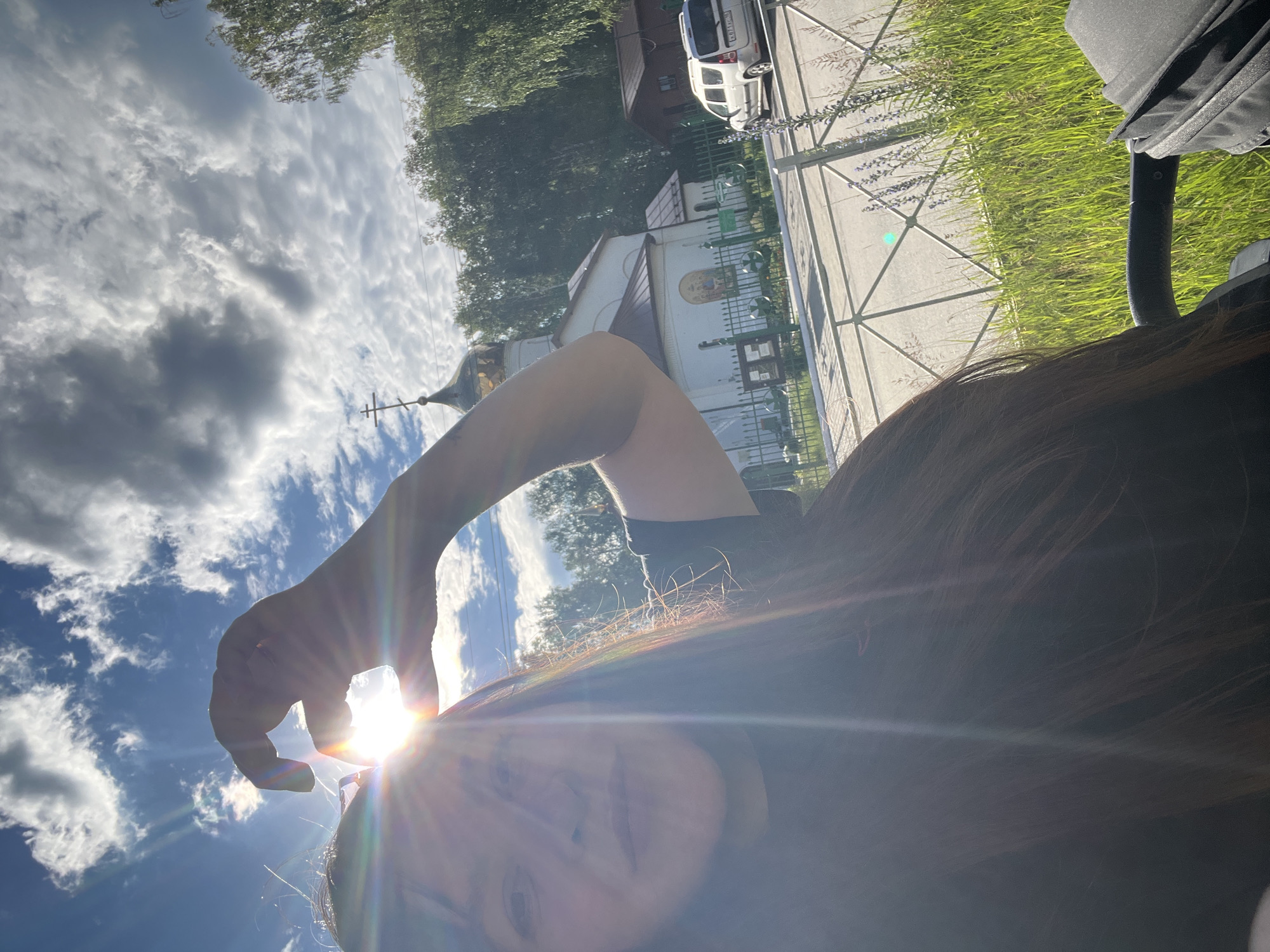 A young woman with long, dark hair is taking a selfie outdoors on a sunny day. She is partially obscuring the sun with her hand, creating a lens flare effect that illuminates her face and hair. In the background, a white church with a gold dome and a cross is visible, along with trees, a fence, a road, and a silver van. The image appears to have been taken in Noviy, Russia, given the architecture and vegetation.