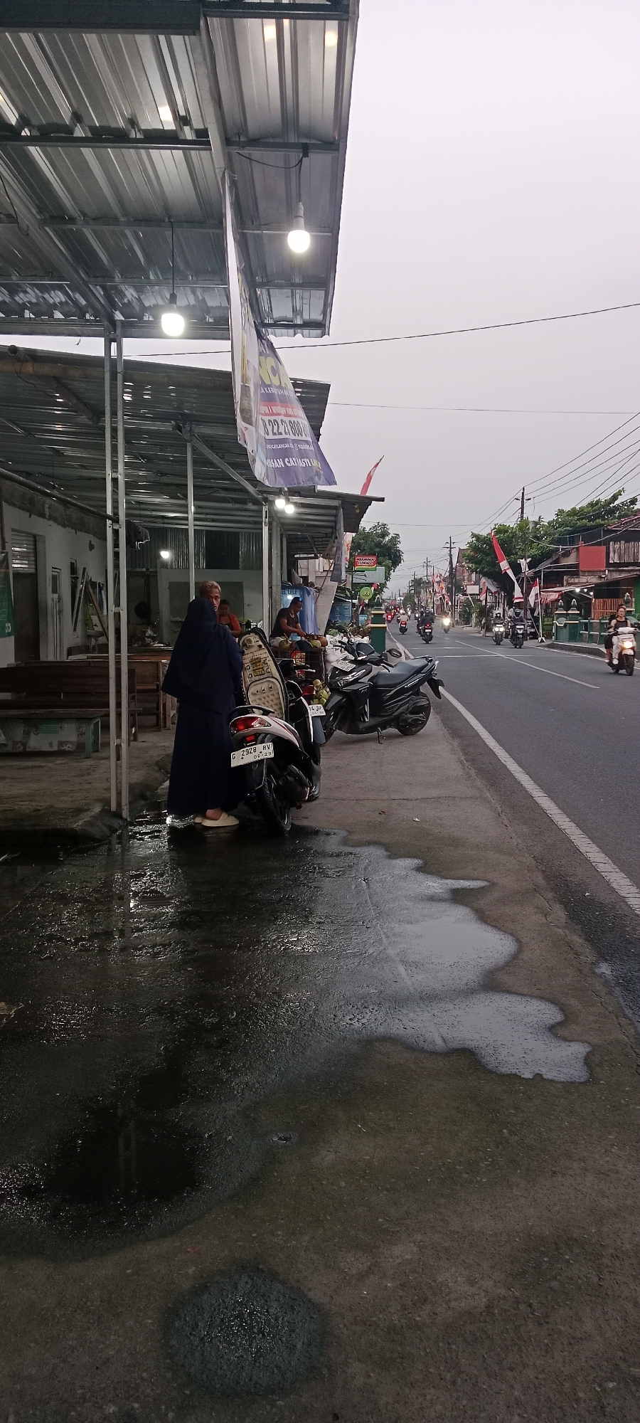 A street scene in Maguwoharjo, Indonesia, shows a covered outdoor area with two motorcycles parked in front of it. A person wearing a dark blue hijab and white shoes stands beside one of the motorcycles. Water is pooled on the ground in front of the covered area and on the sidewalk. In the background, a road is lined with buildings and trees, with several motorcycles and cars driving on it. Indonesian flags are visible on poles along the street. The sky is overcast.