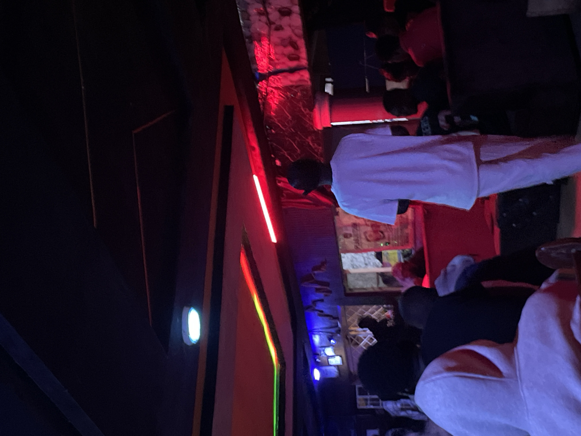 The image depicts an indoor scene, likely a nightclub or bar in Durumi, Nigeria, characterized by low lighting and vibrant neon accents. The ceiling features a dark paneling with embedded lights, including a prominent circular white light and strips of red and green neon along the edges of a lowered section. A man in a white t-shirt and pants, wearing a black cap, stands in the center of the frame, facing away from the camera. He appears to be in a relatively open area of the establishment. In the foreground and to the left, several people are visible from behind, their features obscured by darkness and the angle. They are seated or standing, suggesting a gathering or event. The background shows a wall with some decorative elements, potentially posters or textured surfaces, illuminated by blue and red lights, contributing to the ambient atmosphere of the venue. The overall impression is one of a lively nighttime environment.