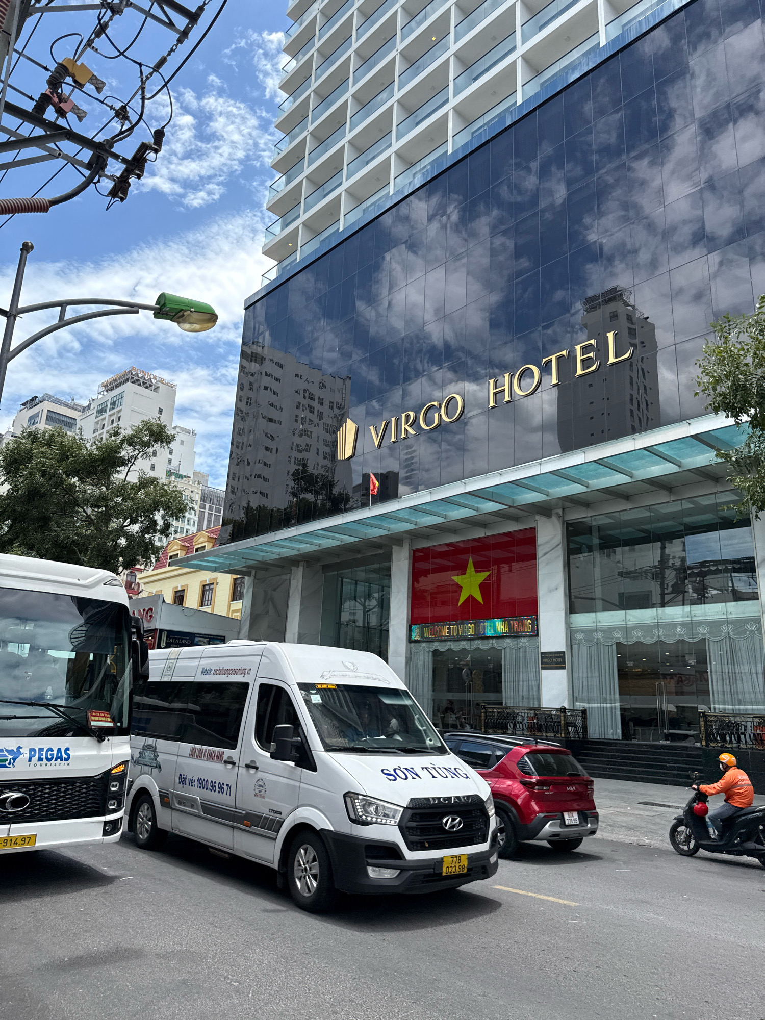 The image depicts a street scene in Nha Trang, Vietnam, featuring the facade of the Virgo Hotel. The hotel is a tall, modern building with a dark, reflective glass exterior. Prominently displayed on the building is the hotel's name, "VIRGO HOTEL," in gold lettering. Below the name, the Vietnamese flag is visible, along with a sign that reads "WELCOME TO VIRGO HOTEL NHA TRANG."
In the foreground, several vehicles are present. A white tour bus, with "PEGAS TOURISTIK" visible on its front, is parked on the left. Next to it is another white van with Vietnamese text and a phone number. A red Kia car is parked behind the vans. A person on a black scooter, wearing an orange jacket and helmet, is riding down the street on the right side of the image.
The environment includes a street with road markings and a sidewalk. Overhead, electrical wires and a street lamp are visible against a blue sky with scattered clouds. Other buildings, including high-rises, can be seen in the background. The overall scene suggests a busy urban environment on a daytime.