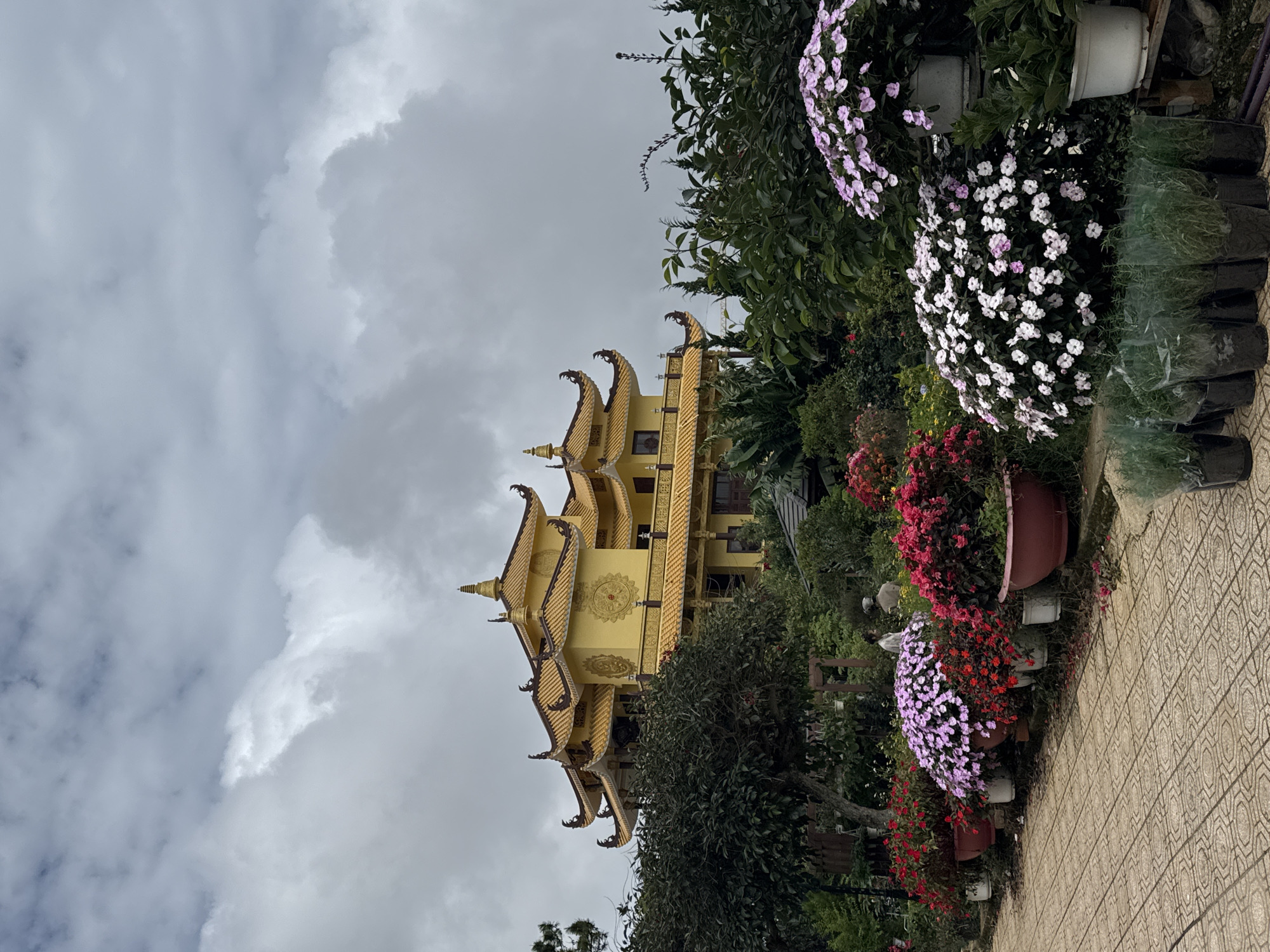 A Buddhist temple with a bright yellow, multi-tiered roof is visible in the background, set against a cloudy sky. In the foreground, a paved walkway leads past several large planters filled with vibrant pink, red, and white flowers. Lush green foliage and trees surround the temple and the flower arrangements. Two individuals are partially visible in the mid-ground, walking near the planters. The scene appears to be an outdoor garden area, possibly part of the Truc Lam Zen Monastery in Da Lat, Vietnam.