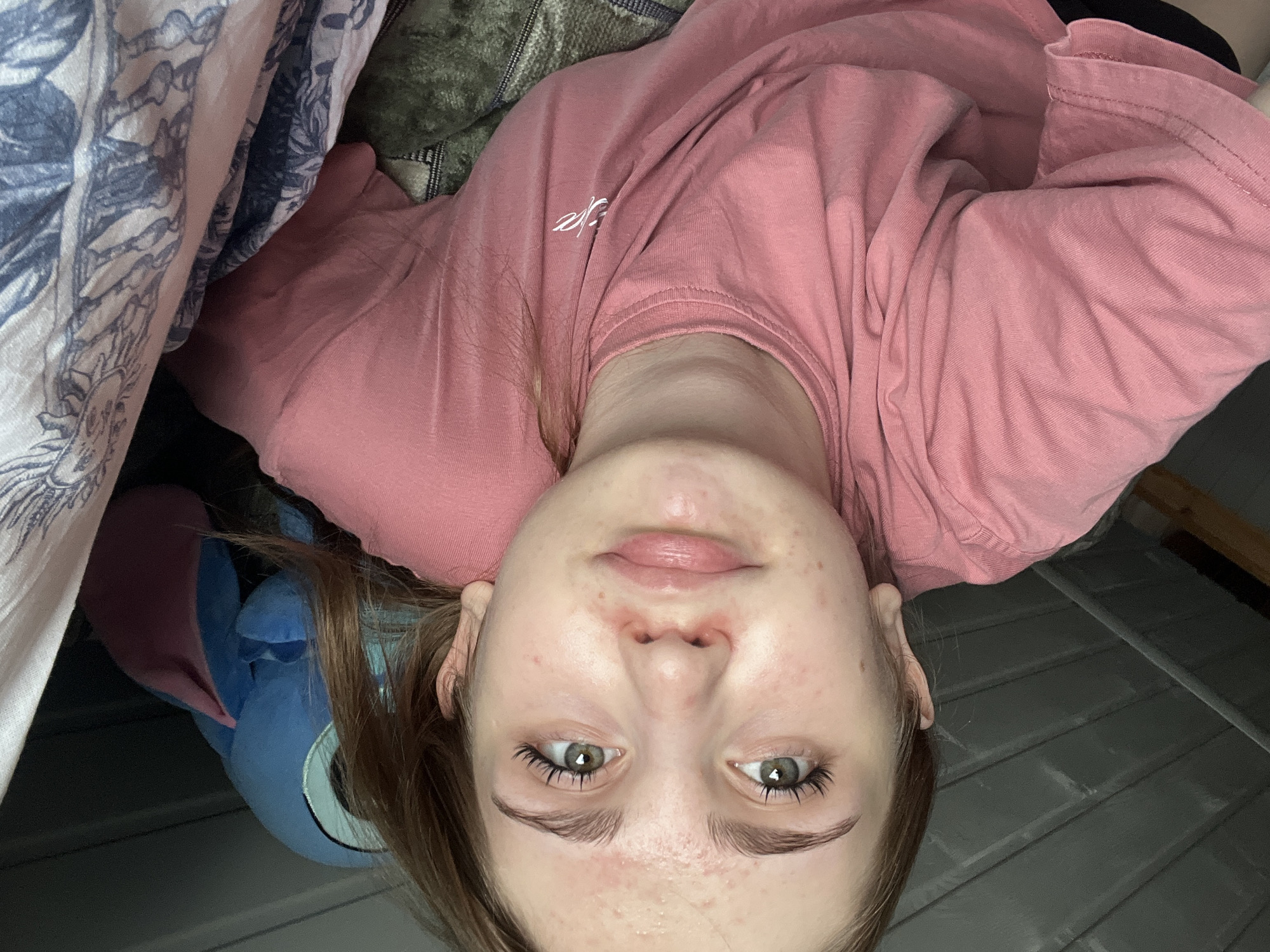 The image shows a young person with fair skin, brown hair, and green eyes, looking directly at the camera. They are wearing a pink t-shirt and are lying down, partially covered by blankets. A blue stuffed toy is visible behind their head. The background consists of a grey wall with horizontal lines and a glimpse of a wooden surface. The scene appears to be indoors, possibly in a bedroom. The image was taken in the city of Ashkadar, Russia.
