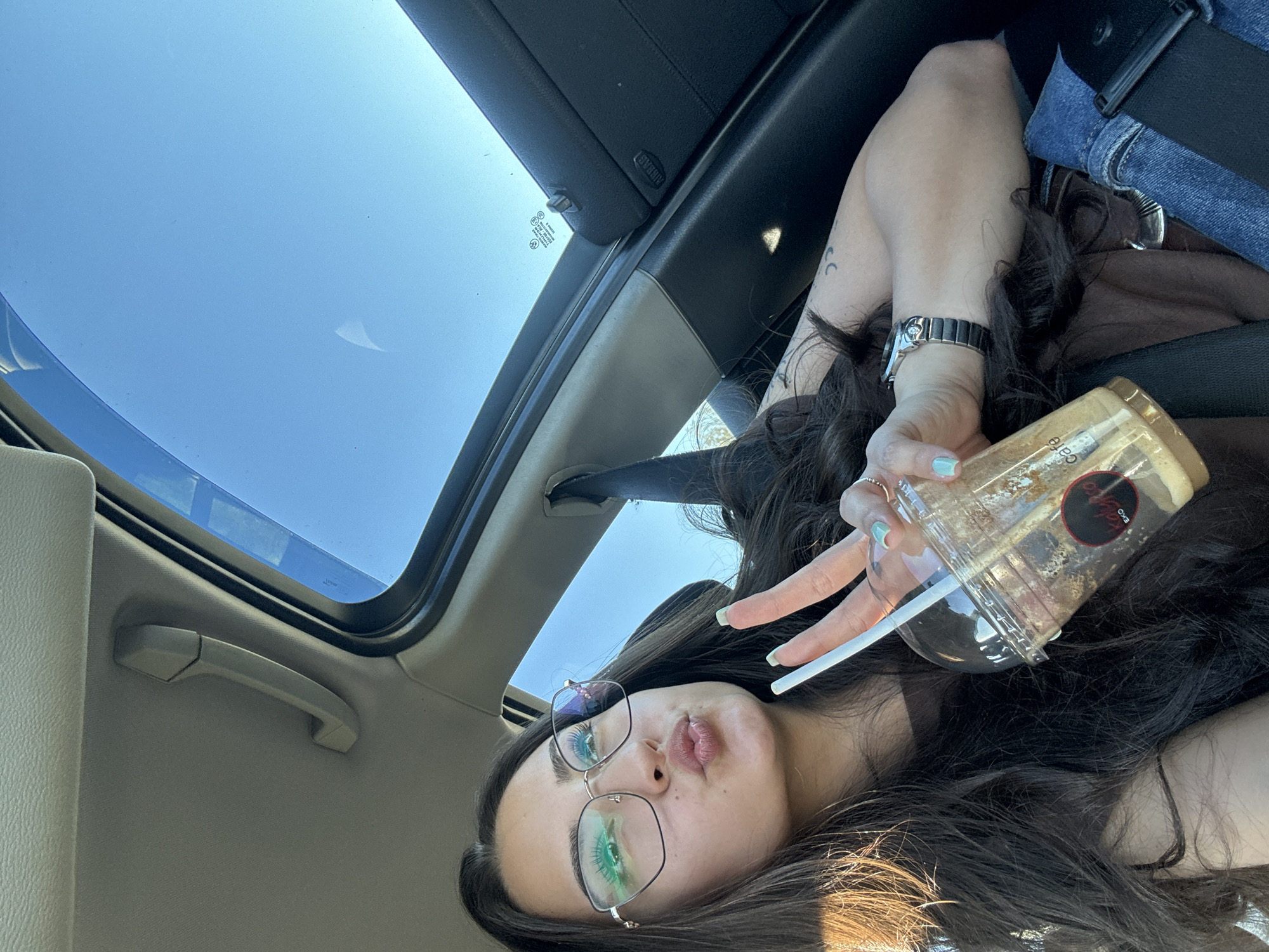 A woman is photographed from a low angle while inside a car with an open sunroof. The sky is visible through the sunroof. The woman has long, dark hair, wears glasses, and holds a plastic cup with a straw in her right hand. She is wearing a watch on her left wrist. The car's interior is visible. There is no information available to determine location as Piperevo, Bulgaria.