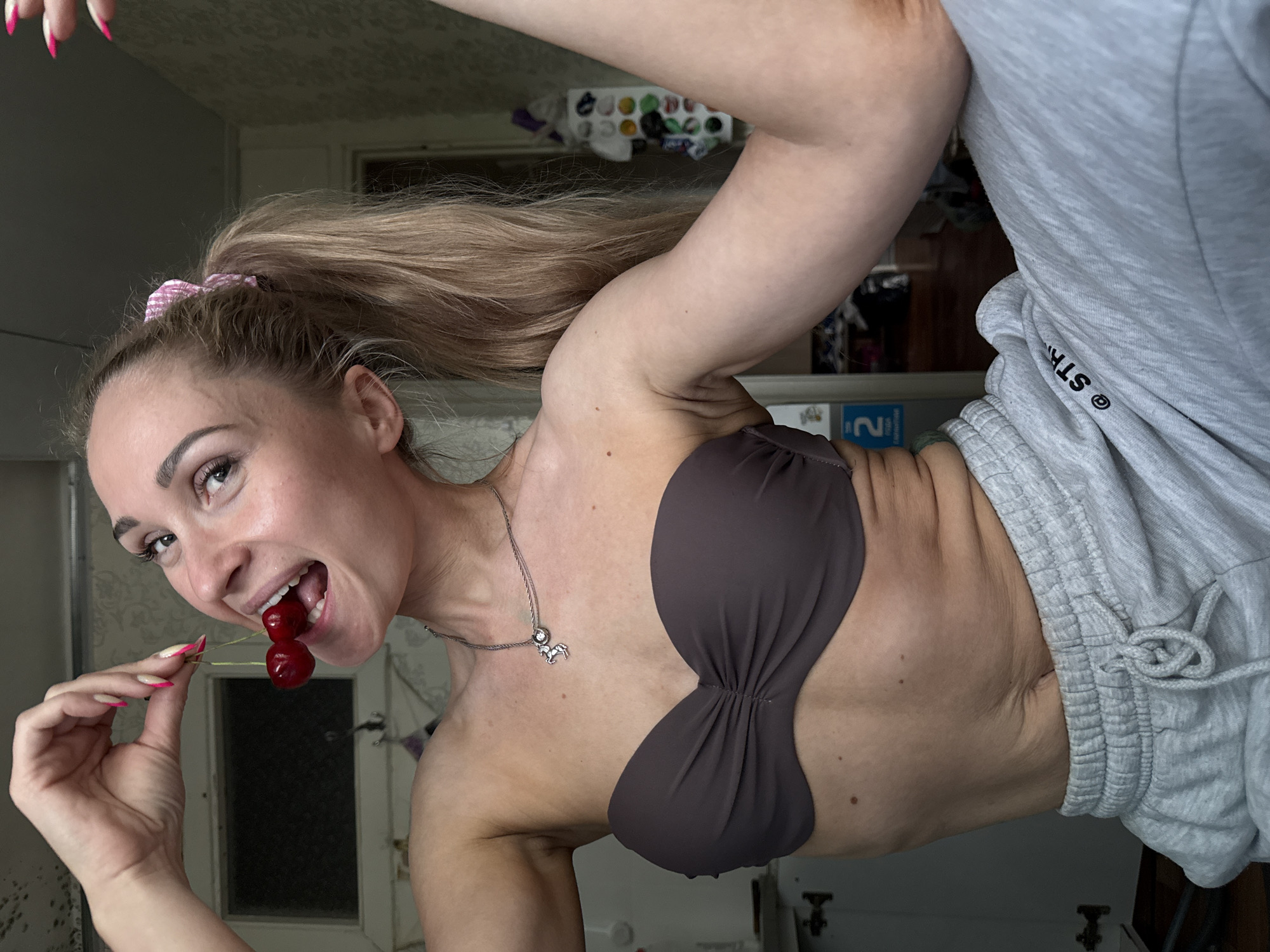 A woman is seen from the waist up. She has long blond hair pulled into a ponytail with a pink scrunchie. She is wearing a brown bra and gray sweatpants. She is holding two cherries by the stems and placing them in her mouth. The background is blurry, but appears to be a room inside a home. The location is Chaikovsky, Russia.