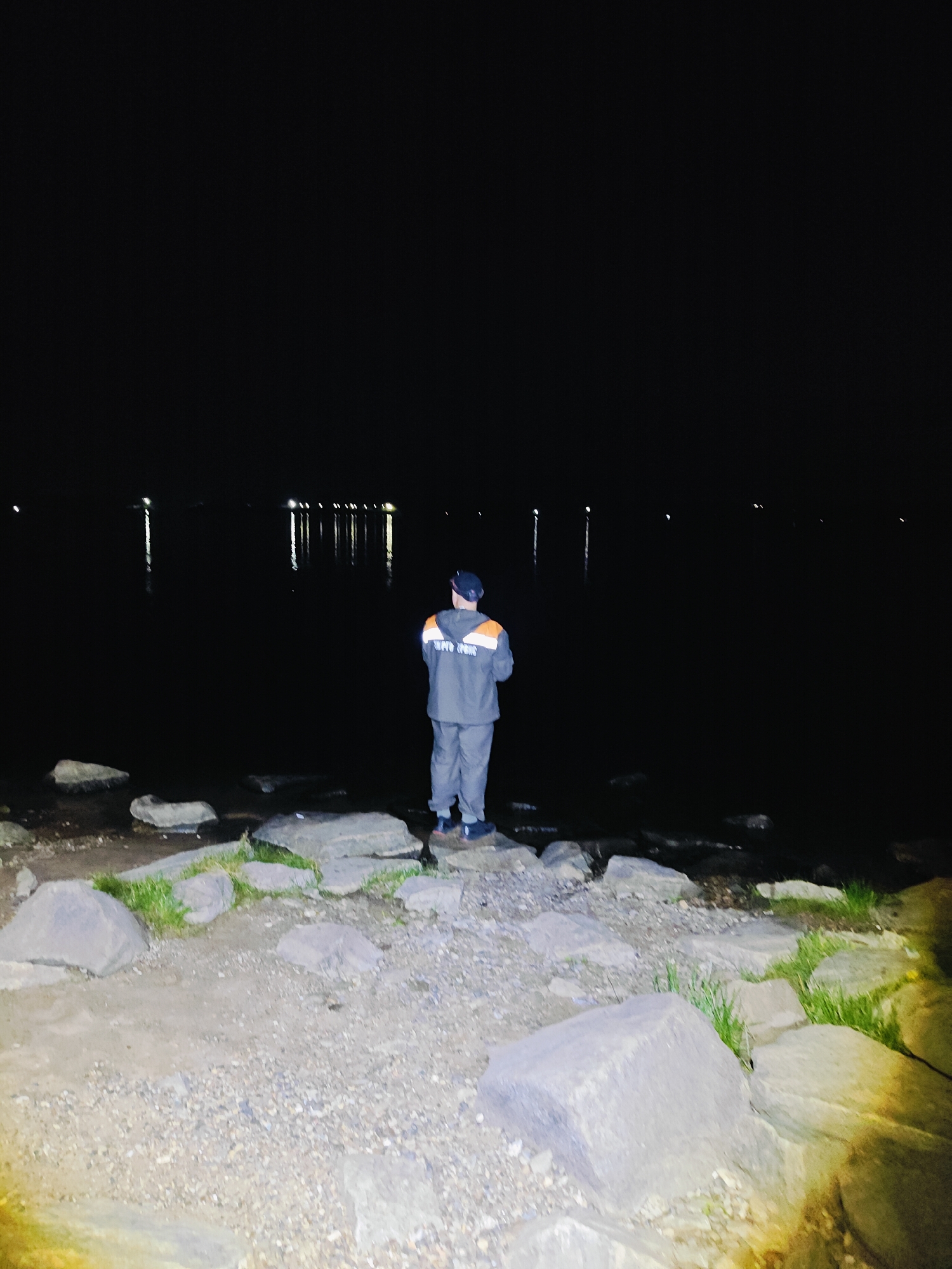 A person in work clothes stands on the shore of a body of water at night. The person is wearing a dark jacket with orange accents and matching pants, and a dark cap. They are facing away from the camera, looking out at the water. Several lights are visible in the distance across the water, possibly streetlights or lights from a building. The shore is rocky, with large stones and gravel. There are patches of grass growing between the rocks. The water is dark and still, reflecting the distant lights. The sky is black, indicating it is nighttime. The location is the shore of the Kama River in Chaikovsky, Russia.