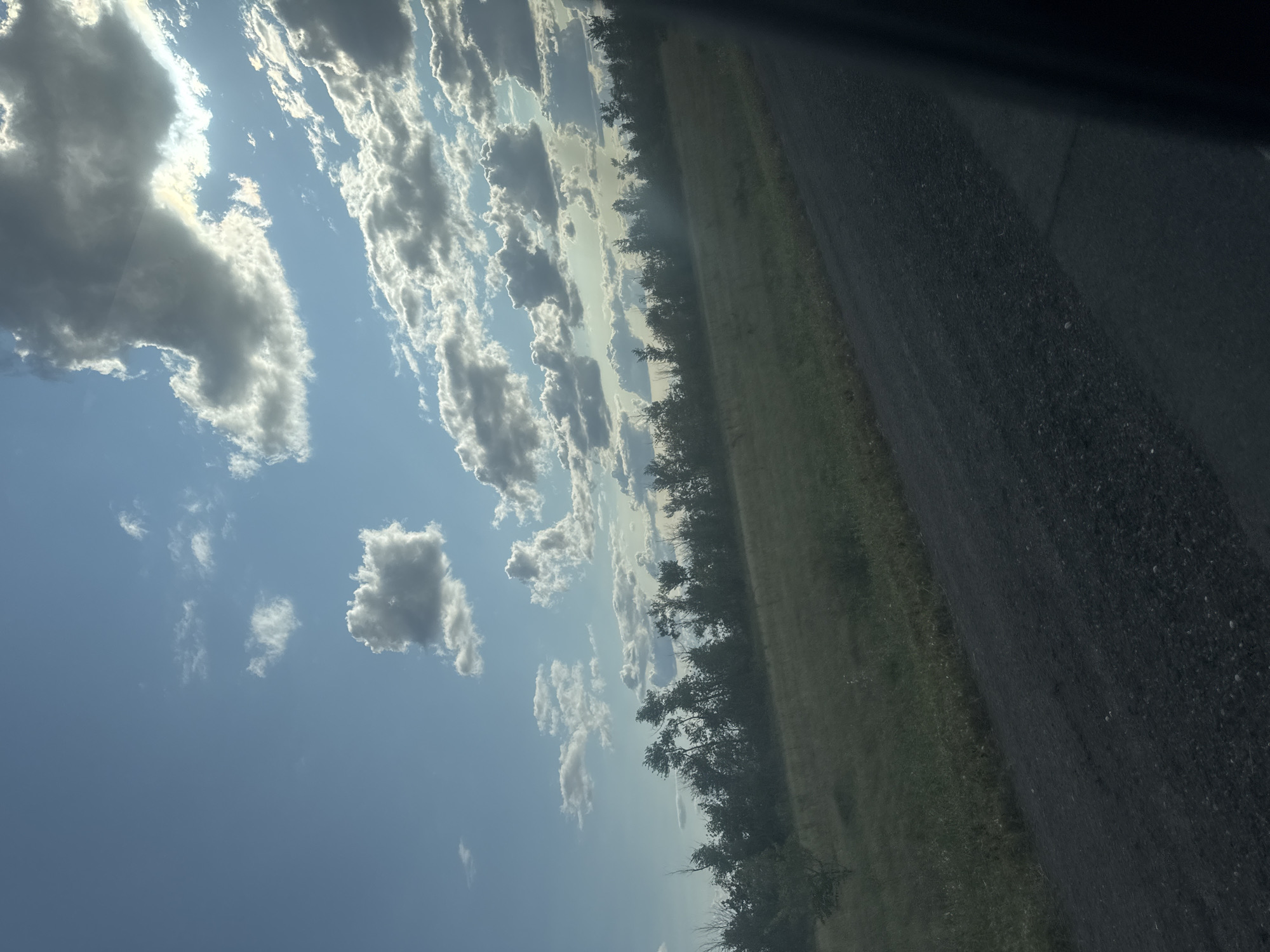 The image depicts a rural landscape under a partly cloudy sky. A dirt road runs diagonally across the lower right portion of the frame, bordered by a field of dry grass. In the distance, a line of trees forms a dense hedgerow against the horizon. The sky is a vibrant blue, with scattered cumulus clouds. The sun is visible, casting a bright glare through a break in the clouds, illuminating parts of the sky and the tops of the trees. The scene is devoid of any people or specific landmarks that would indicate its precise location within Perevolotsky, Russia. The overall impression is one of an open, natural environment.