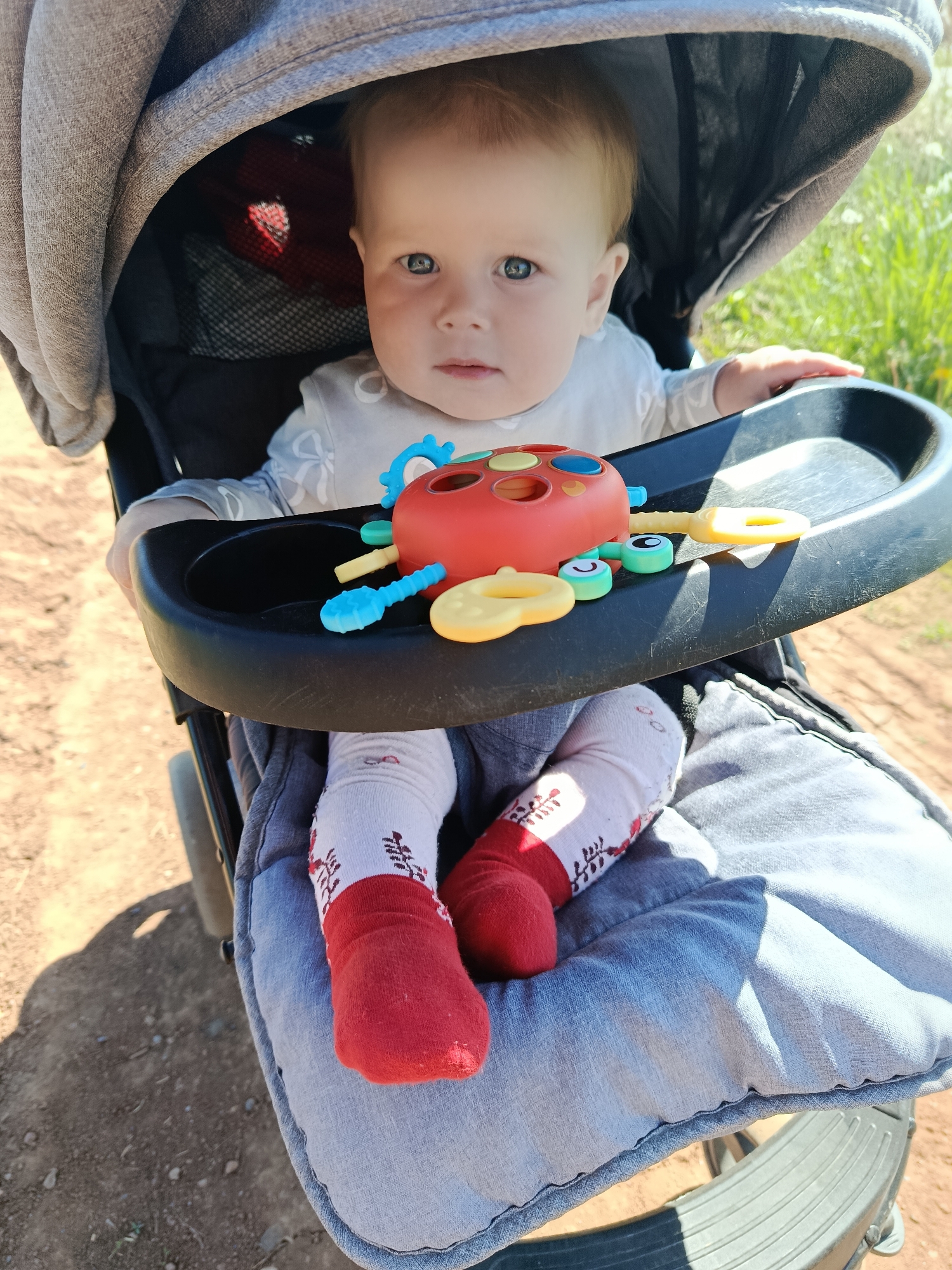 The image shows a baby sitting in a stroller outdoors. The baby has fair skin, light hair, and blue eyes, and is wearing a long-sleeved shirt. The baby's legs are clad in white socks with a red toe and a dark, decorative pattern near the ankle. The stroller has a gray fabric hood partially covering the baby. A black tray is attached to the stroller in front of the baby, and on this tray sits a colorful toy with various shapes and holes. The seat of the stroller is upholstered with a light blue fabric. The ground visible outside the stroller is dirt. The location is not discernible as Фоки, Russia, from the information in the image.