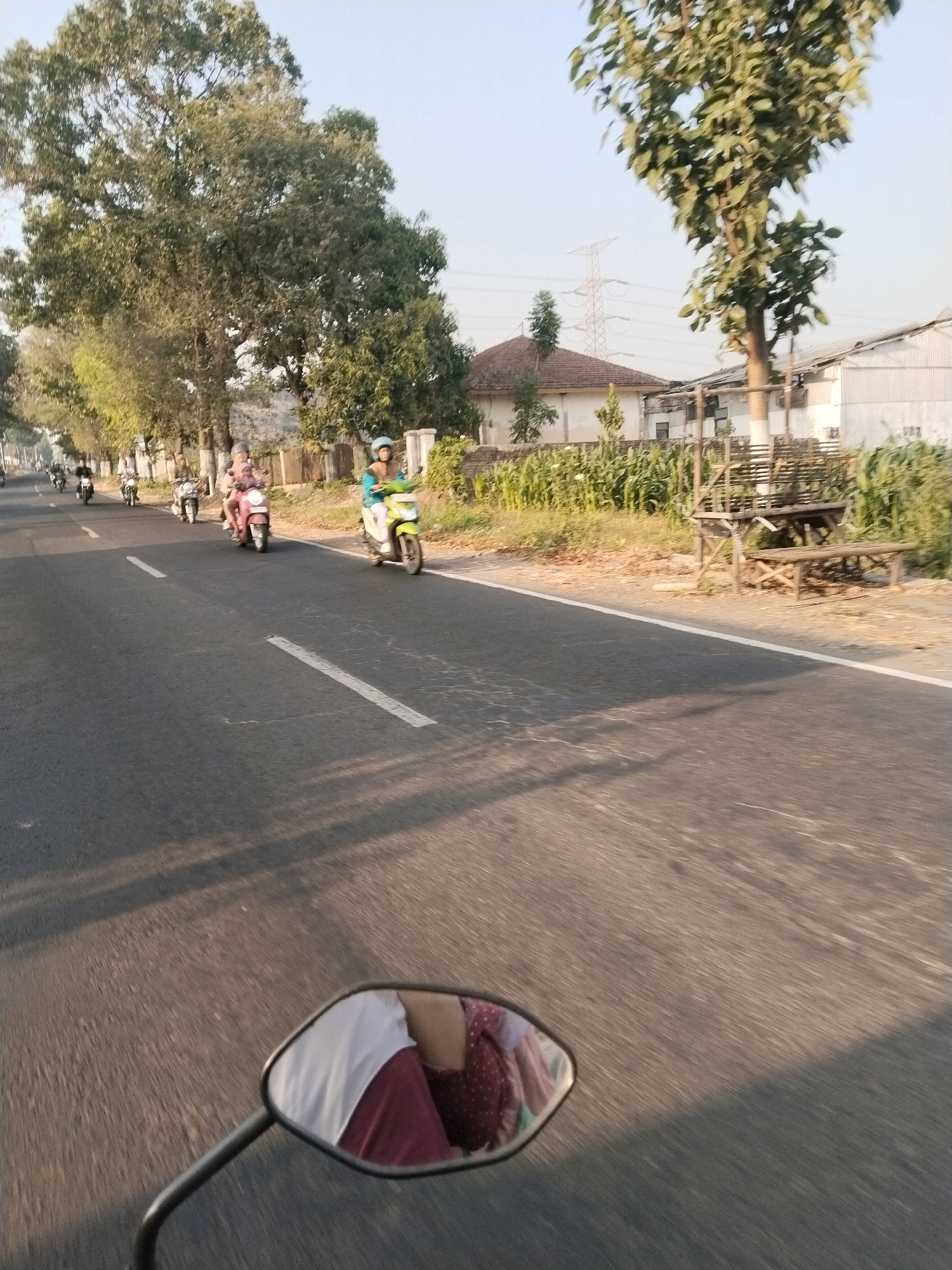 In Simbaringin, Indonesia, a first-person perspective shot captures a view of a road with several individuals on motorcycles. The road is paved with white lines marking lanes and is bordered by trees and vegetation. In the background, buildings and a transmission tower are visible. In the foreground, a motorcycle's side mirror reflects the upper torso of a person wearing a white shirt and a burgundy patterned top, suggesting the photographer is riding a motorcycle. The overall atmosphere is sunny with a clear sky.