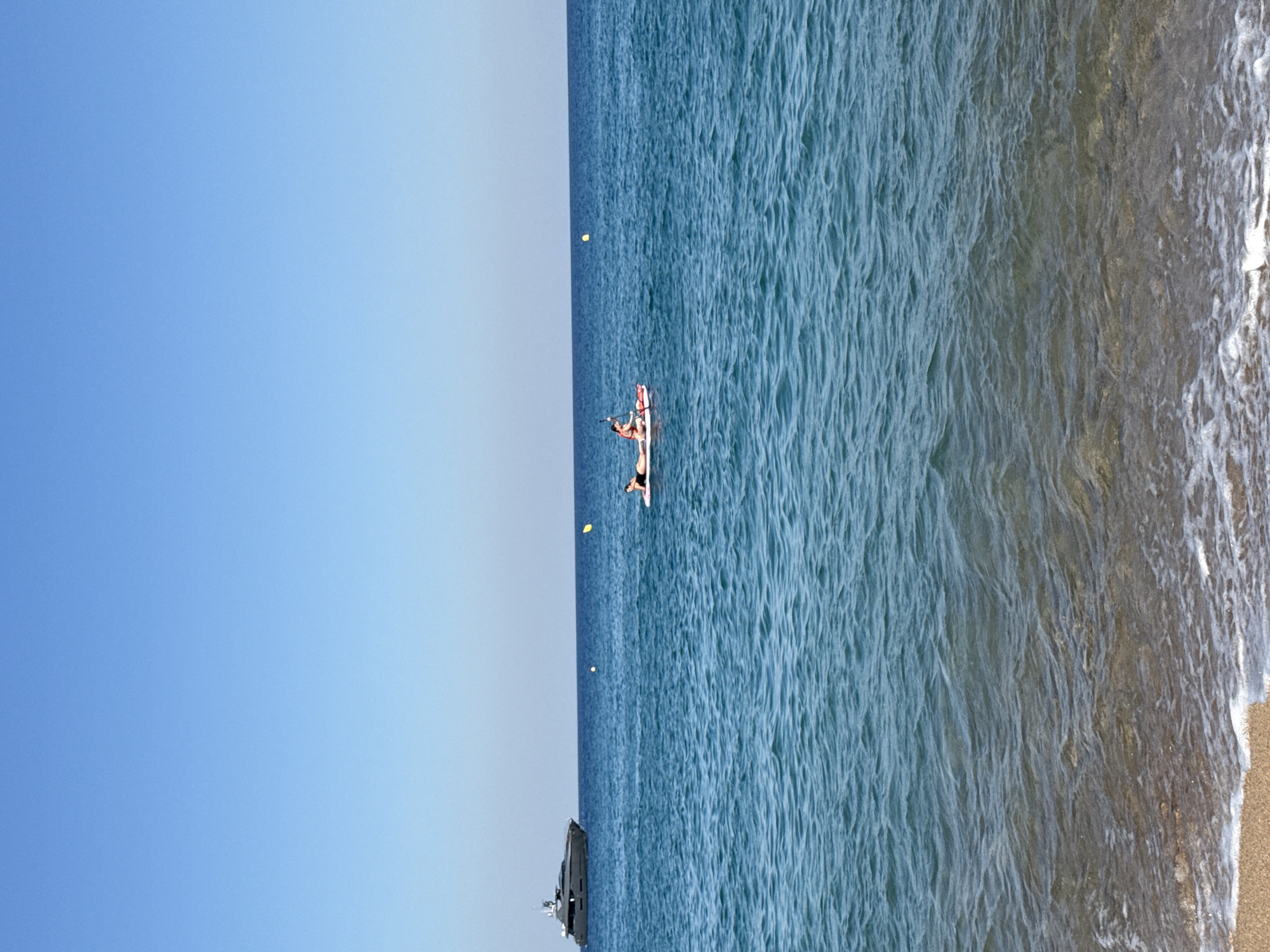 In the waters off the coast of Blanes, Spain, under a clear blue sky, a large, dark yacht is anchored in the distance to the left of the frame. In the mid-ground, a person is paddleboarding, lying on the board with their arms outstretched. A small, pink and white paddleboard is visible. Two yellow buoys are present in the water between the yacht and the paddleboarder. The foreground shows the gentle waves of the Mediterranean Sea lapping onto a sandy shore.