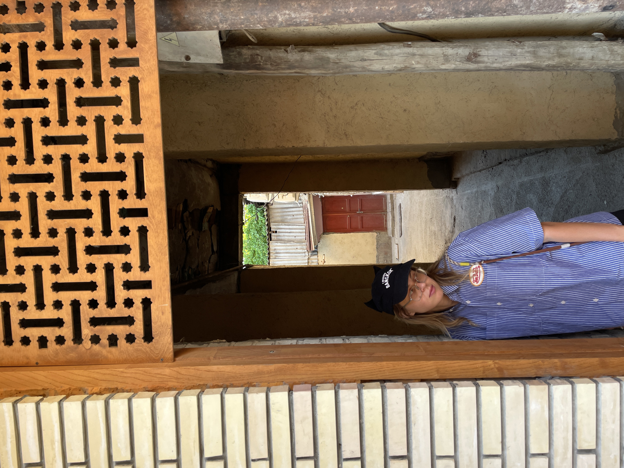 The image shows a person wearing a blue and white striped shirt, a black baseball cap with "Bangkok" written on it, and glasses, standing in what appears to be a narrow passageway or alley. The passageway is constructed of brick and wood, and there is a wooden lattice panel on the left. At the end of the passageway, a red door is visible, and there are some plants and fencing. Based on the building style and the door, this could be a traditional area in Tashkent, Uzbekistan.