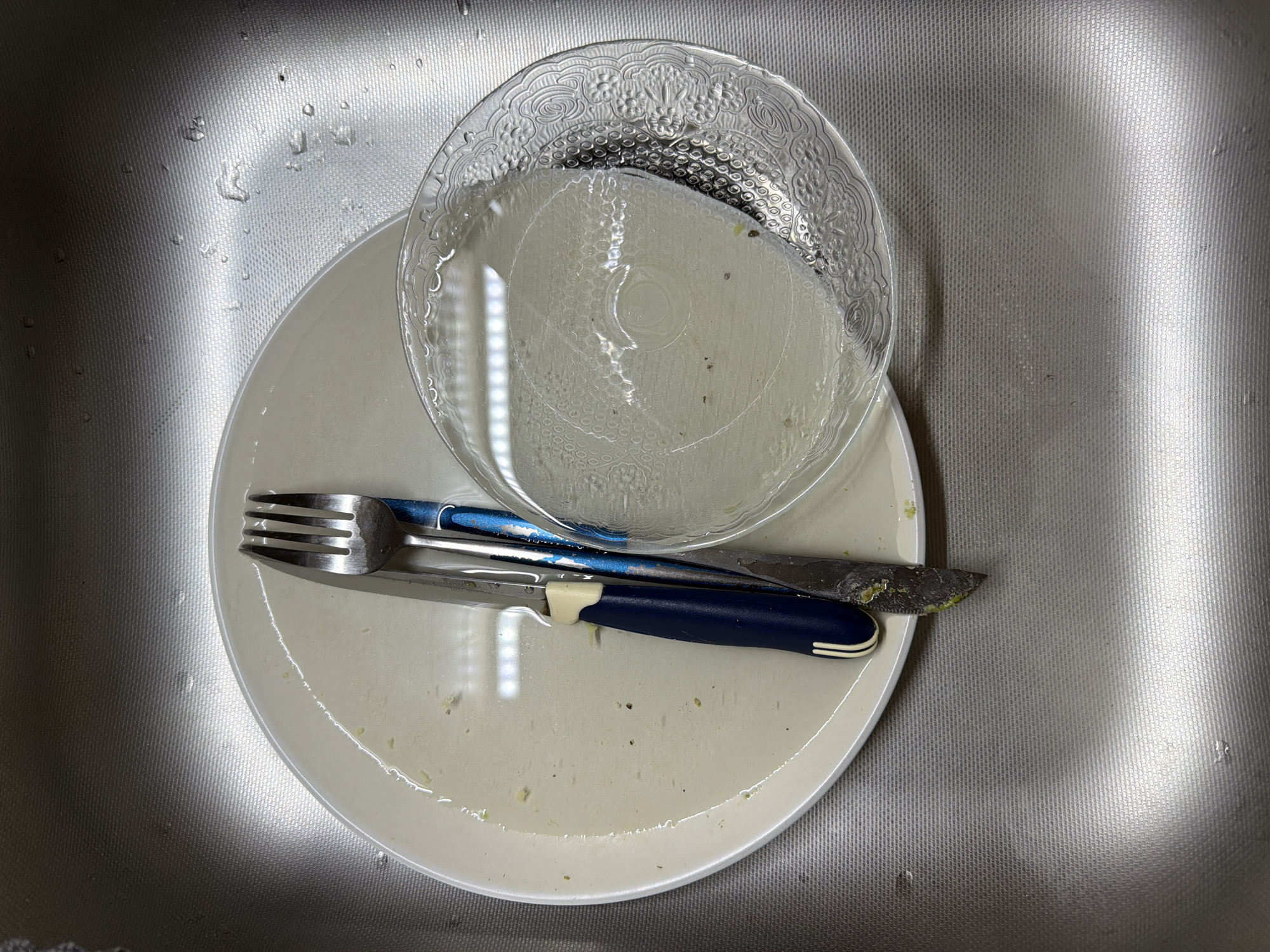 A white plate sits in a sink filled with murky water. On the plate are a fork, a knife, and a small glass bowl. The fork and knife are resting on the plate, and the knife has some green residue on its blade. The glass bowl contains some water and appears to have a decorative edge. The sink is made of stainless steel and shows signs of use, with water droplets and a textured surface. The overall scene suggests dishes that have been placed in the sink for washing. The image is taken from a top-down perspective.