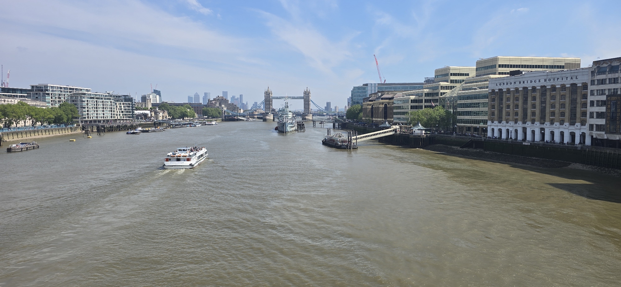 This is an eye-level panoramic view of the River Thames in London, United Kingdom. In the background, Tower Bridge is seen spanning the river, connecting the city center. A white passenger boat, filled with tourists, is moving along the river in the direction of the viewer. Several ships and boats of varying sizes are docked along the river, and the foreshore is visible along the banks. Various buildings and structures line both sides of the river, including the London Bridge Hospital on the right bank. The sky is light blue with scattered clouds.