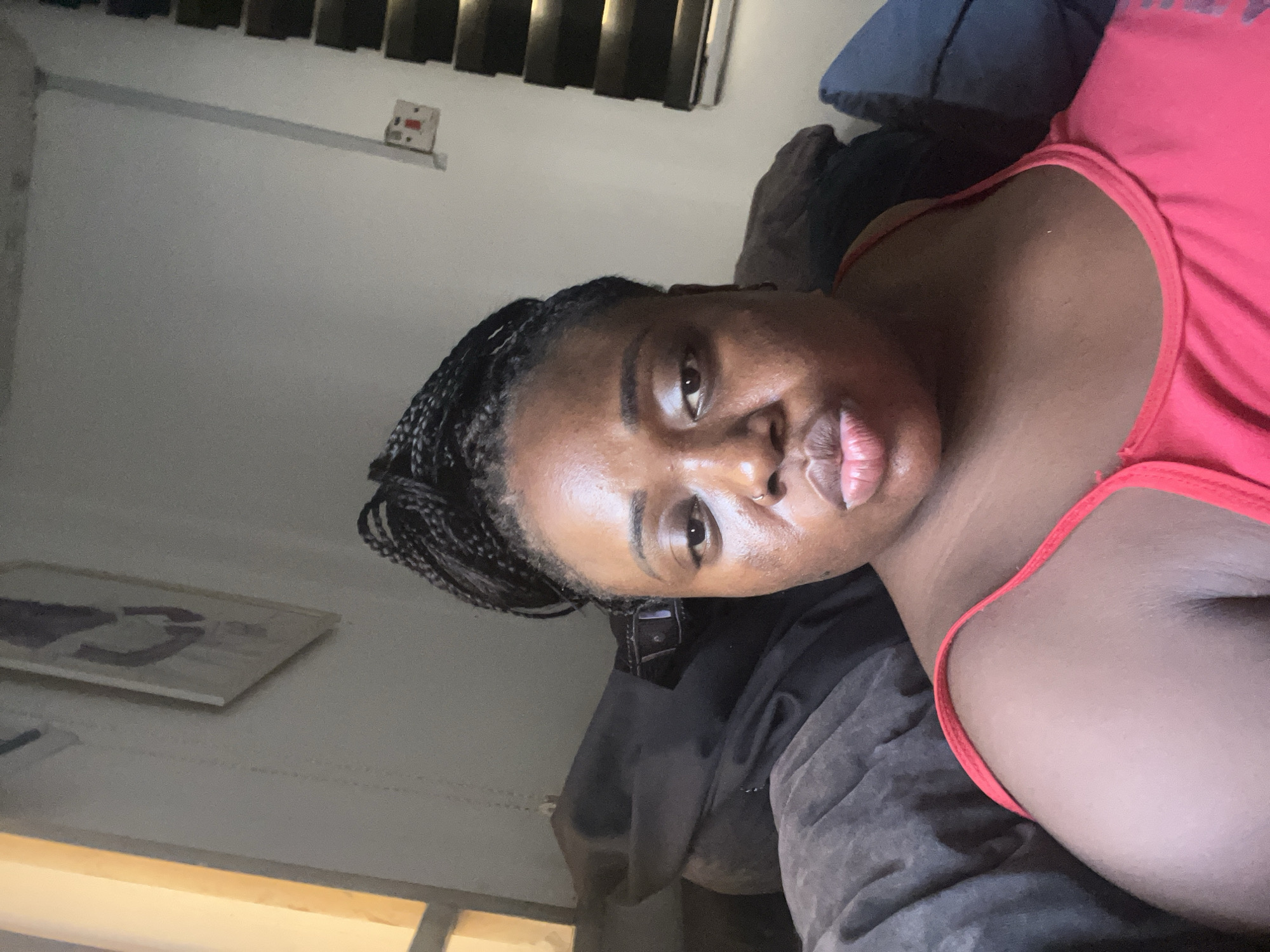 A woman with dark skin and braided hair is lying on a gray upholstered surface, possibly a couch. She is wearing a red sleeveless top. The background shows a wall with blinds and an electrical outlet. A framed picture hangs on the wall. The location is likely a residential interior. There's no information to assume it's in Abuja, Nigeria.
