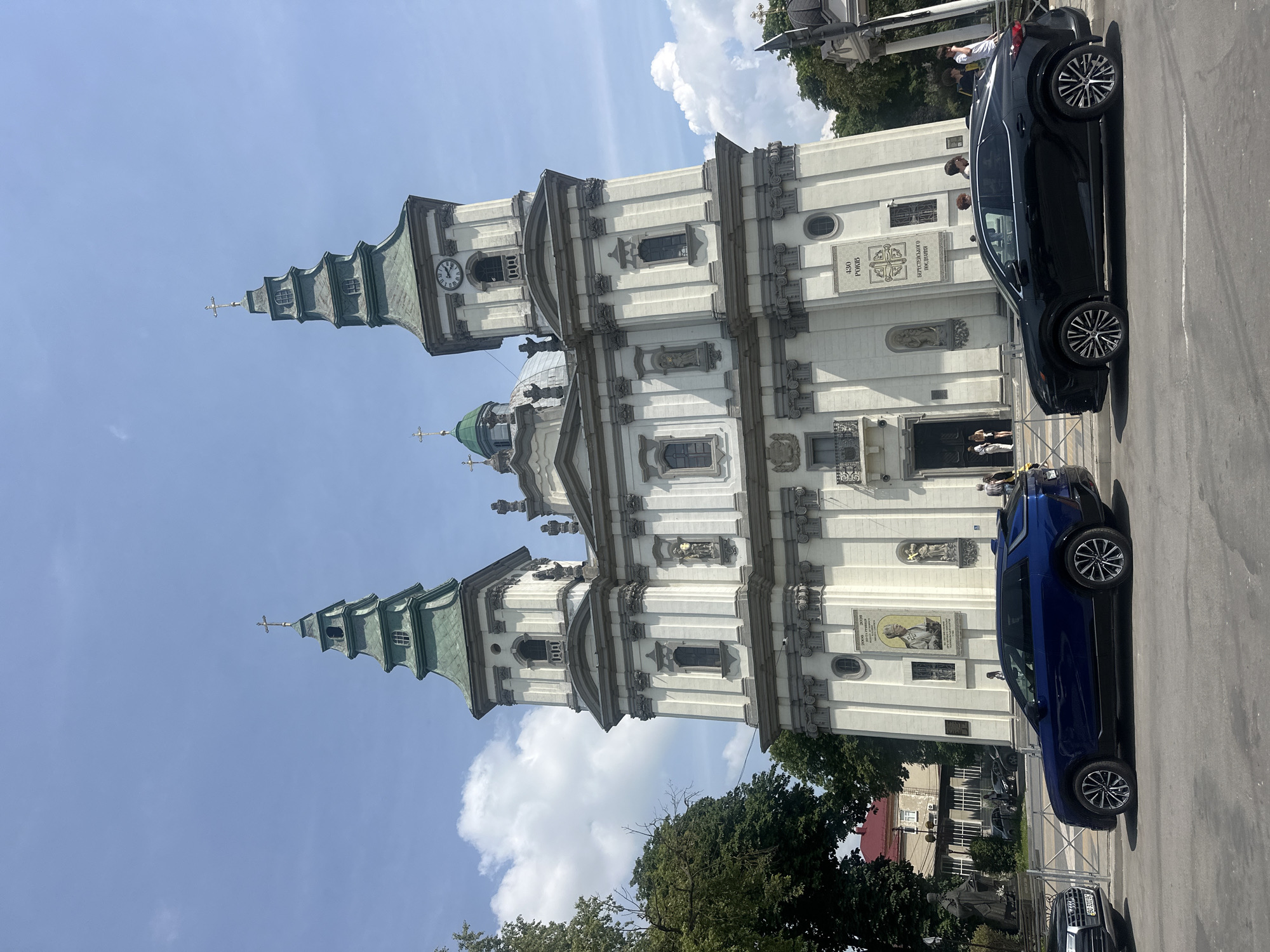 This image shows the Ternopil Cathedral in Ternopil, Ukraine, on a sunny day with a partly cloudy sky. In the foreground, two cars are parked on the street: a dark blue SUV on the left and a black SUV on the right. The cathedral, a large white building with two green-topped towers, is the main subject. The towers have crosses at their peaks, and one tower has a clock. The facade of the cathedral features architectural details, sculptures, and large banners with text. Several people are visible near the entrance of the cathedral and on the sidewalk. Trees and other buildings are present in the background, and a fence separates the sidewalk from the street.
