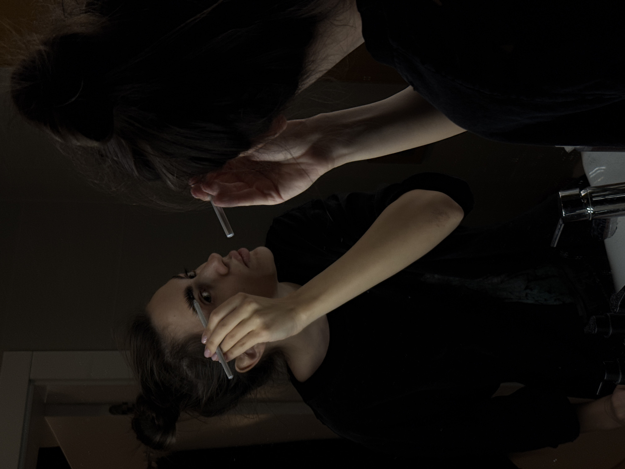 A low-angle shot captures a woman grooming her eyebrows in a bathroom mirror. She is using a metal tool, possibly tweezers or an eyebrow brush, on her right eyebrow. Her hair is pulled back in a low bun. The lighting is dim, with much of the upper portion of the mirror obscured by darkness, possibly reflecting the ceiling. A section of the sink, including the faucet, is visible in the lower right. The location is Unknown, Unknown.