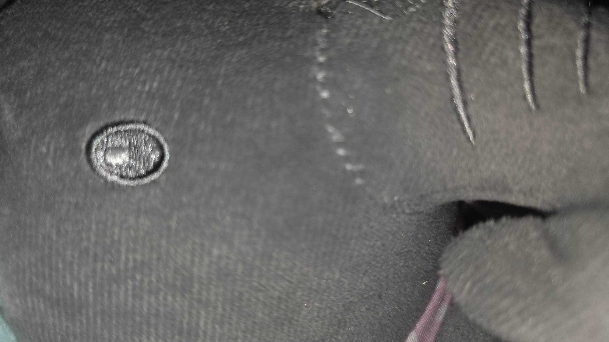 This is a very close-up, out-of-focus image depicting various textures and details on a dark, likely fabric, surface. The primary subjects are abstract elements rather than distinct objects or people.
In the mid-left section, there is a prominent oval-shaped element, which appears to be embroidered or raised from the surface. Within this oval, a faint, indistinct shape, possibly resembling a small animal, symbol, or logo, is visible, though blurred.
Towards the right and upper right, several parallel or semi-parallel vertical lines are discernible. These lines appear to be either raised stitching, textured fabric elements, or possibly light scratches on the surface, varying in length and clarity.
In the bottom right corner of the image, a soft, dark, possibly furry or plush material is present, partially obscuring a glimpse of a striped or patterned fabric underneath, which shows hints of purple or similar dark colors.
The overall color scheme is predominantly dark, consisting of shades of grey and black, with minimal color variation. The image's shallow depth of field renders most elements blurry, emphasizing texture and light reflections over sharp details. There is no discernible activity or event taking place. No specific location cues are visible within the image itself, nor is there any visible text. The time of day or weather cannot be determined from this close-up perspective.