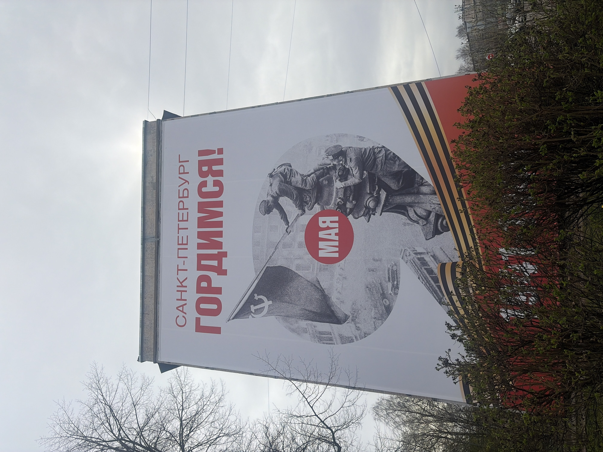 The image shows a large banner in Saint Petersburg, Russia. The banner features the text "САНКТ-ПЕТЕРБУРГ ГОРДИМСЯ!" (Saint Petersburg is Proud!) in red, along with a depiction of Soviet soldiers, a Soviet flag, and the word "МАЯ" (May) in a red circle. The banner also incorporates a Saint George ribbon design. The banner is attached to the side of a building, with trees and a cloudy sky visible in the background.