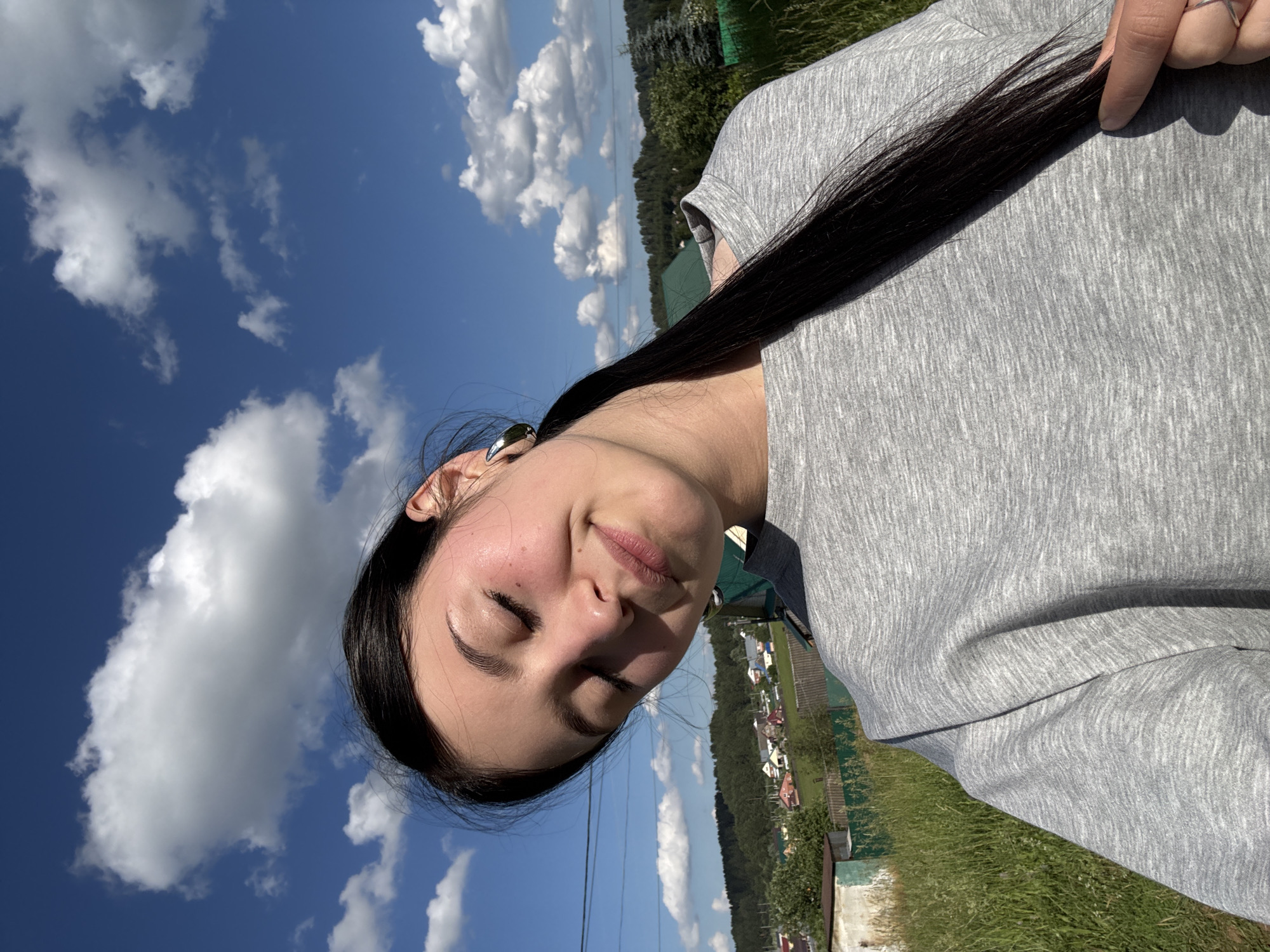 In the city of Fokino, Russia, a young woman with dark hair and closed eyes smiles slightly, tilting her head back. She is wearing a gray sweatshirt and a silver hoop earring in her left ear. Her long, dark hair is pulled back and cascades over her shoulder. The background shows a rural landscape with green grass, trees, houses with red roofs, and a bright blue sky filled with white, fluffy clouds. Power lines are visible in the upper left portion of the image. The woman's right hand is visible near the bottom right of the frame, touching her hair. The lighting suggests it is daytime, likely sunny.