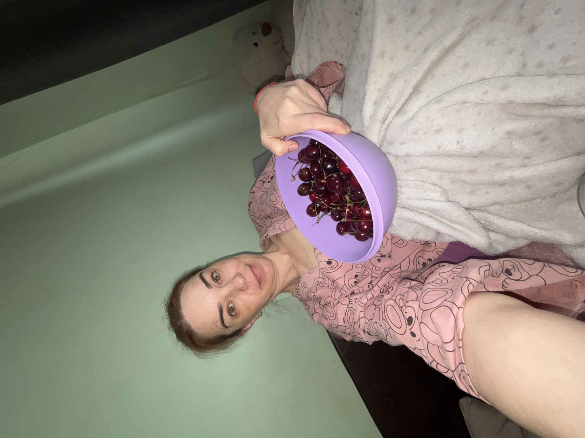 A woman is lying on a bed, holding a bowl of cherries. The room is dimly lit with a light green wall and a dark brown headboard. A white teddy bear is visible in the background. The woman is wearing a pink pajama top with a teddy bear pattern. She has light brown hair and is looking at the camera with a slight smile. The bowl is purple and filled with dark red cherries.