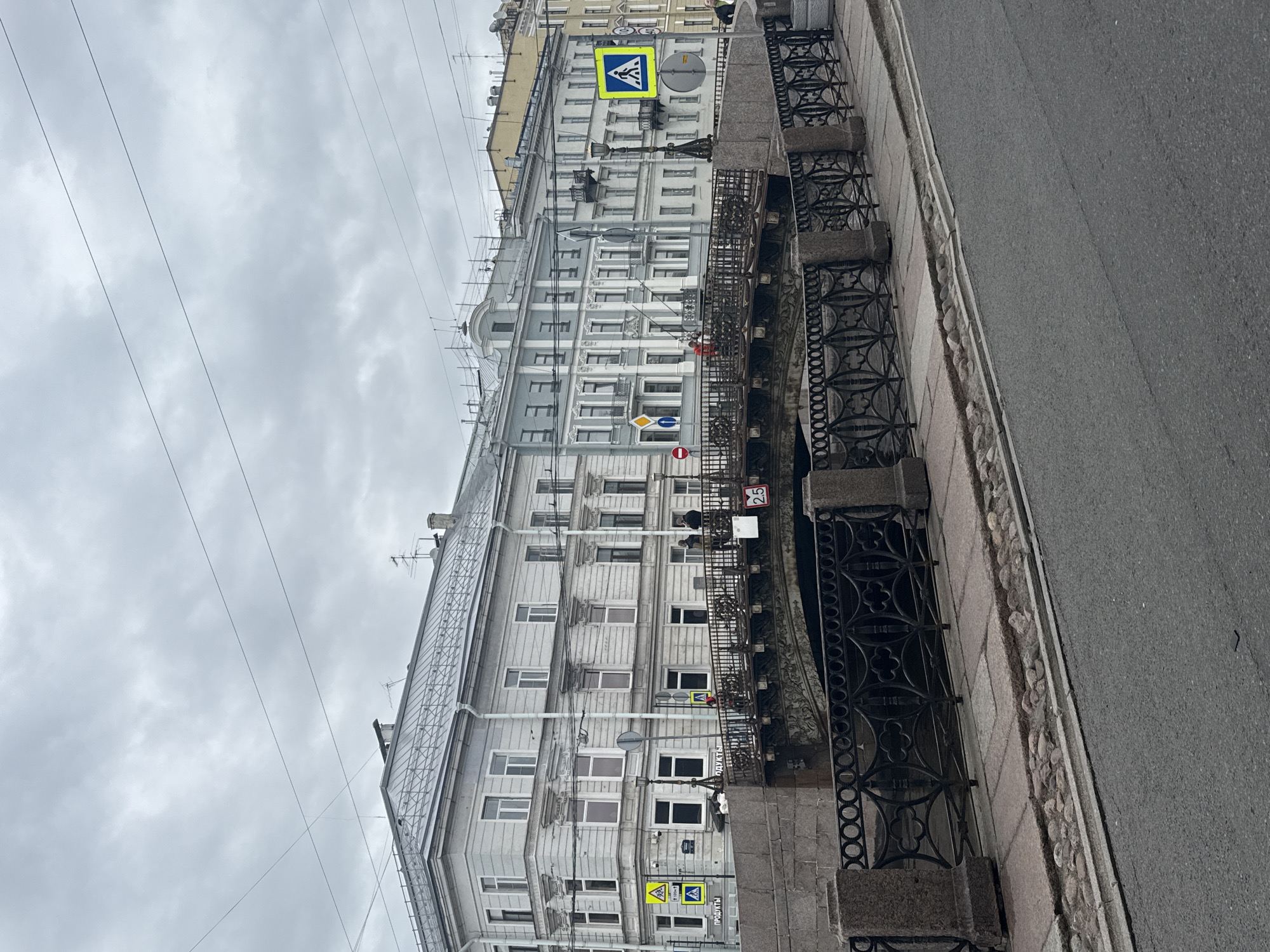 The image depicts a street scene in Saint Petersburg, Russia, showing a canal lined with an ornate black wrought iron fence and a building with multiple windows and several pedestrian crossing signs. The sky is overcast. The street is paved and wet-looking. A few people can be seen on the far side of the canal, and signs for food products are present on the building. Electrical wires run overhead.
