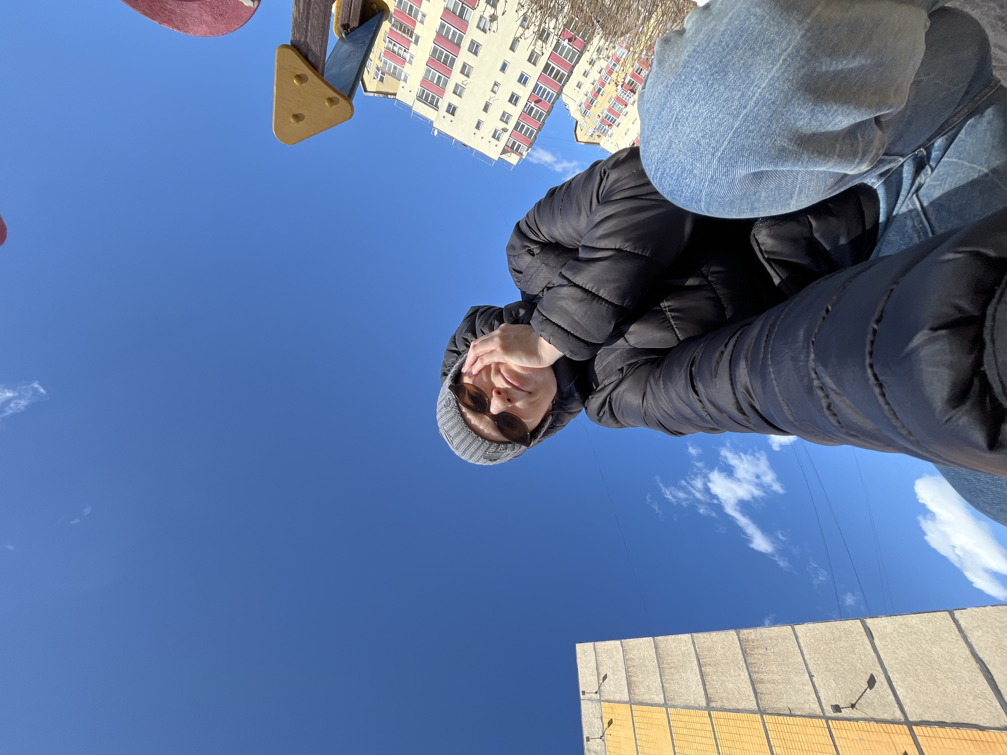 A low-angle, upward-facing photograph shows a person wearing a gray knit hat, sunglasses, and a black puffer jacket, with blue jeans visible. The person appears to be holding the camera, looking down. Behind them is a clear blue sky with a few wispy clouds. A portion of a multi-story building with cream-colored walls and red accents is visible, along with a section of a playground structure. In the bottom of the frame, a light-colored building with rectangular paneling is present. The location is Chaikovsky, Russia.