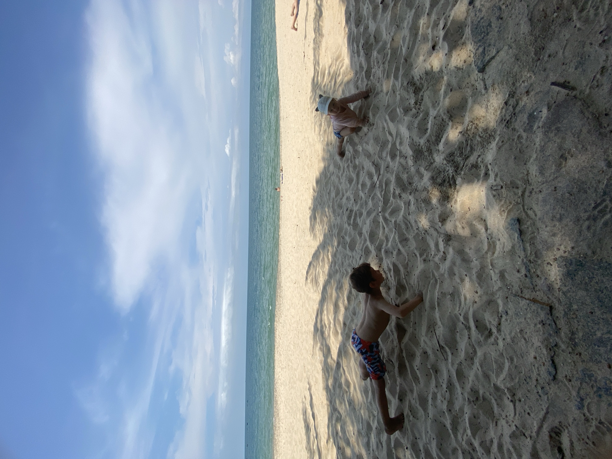 Two young children are on a sandy beach with the ocean and a blue sky with clouds in the background. The child on the left, a boy, is in a push-up position. He is wearing patterned swim trunks. The child on the right, a toddler, is wearing a pink long-sleeved shirt and a light-colored hat with cat ears. They are both on their hands and knees or in a similar crawling or planking position. The sand is textured with shadows cast from unseen palm fronds. In the distance, the water is calm and a few figures can be seen on the beach and in the water. The image appears to have been taken in Phu Thuy Ward, Vietnam, known for its beaches.