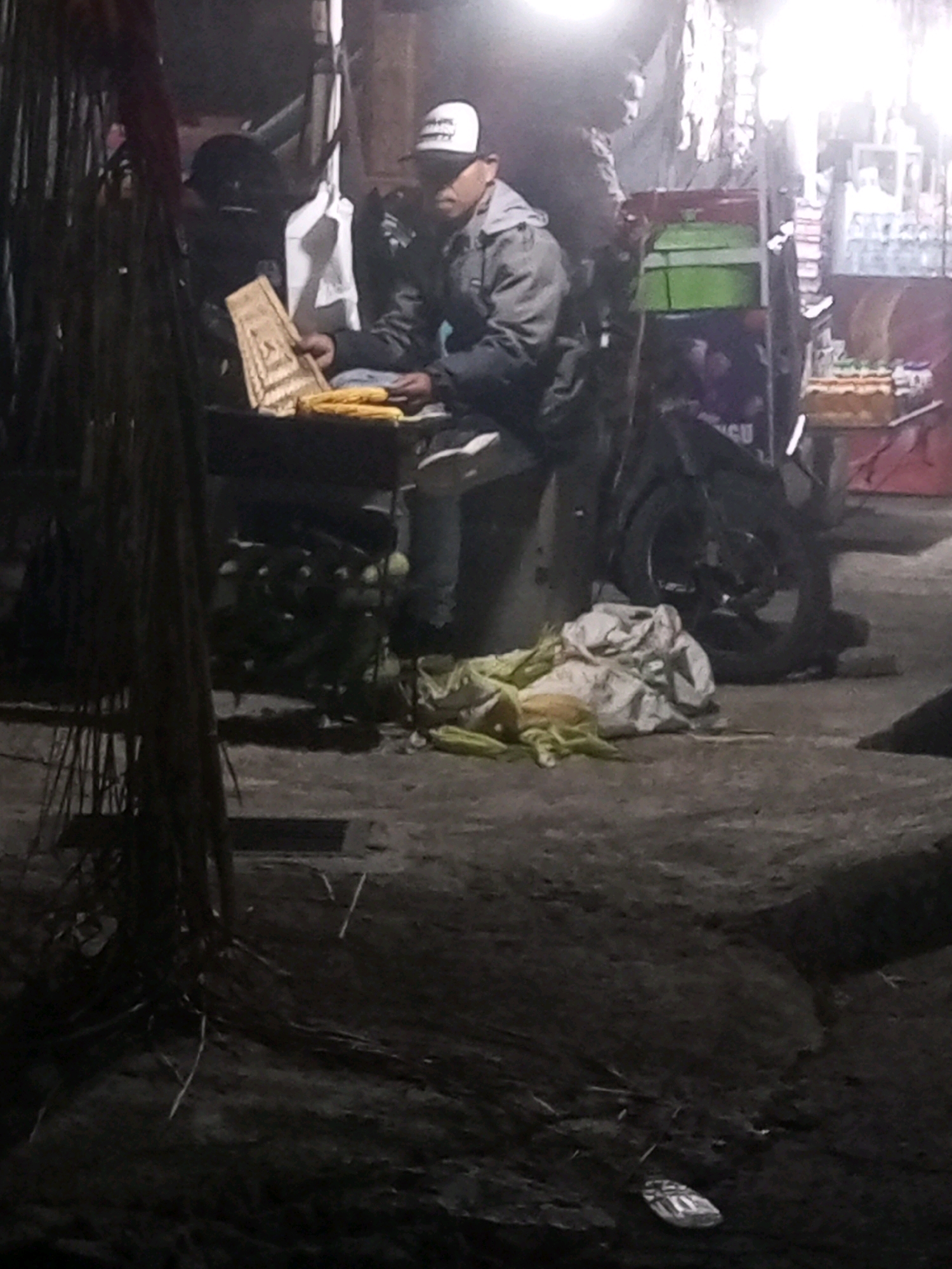 In Keseneng, Indonesia, a nighttime scene unfolds at a street food stall. A person, wearing a cap and jacket, sits behind a table laden with corn. In front of them, a woven basket, possibly used for display or sale, holds more corn. To the right, a motorcycle is parked, and nearby, a bundle of cloth and more corn are scattered on the ground. The stall appears to be illuminated by a bright overhead light, casting shadows and creating a dim ambiance. The surrounding environment is murky, suggesting an urban setting with a gritty, perhaps unpaved, surface.