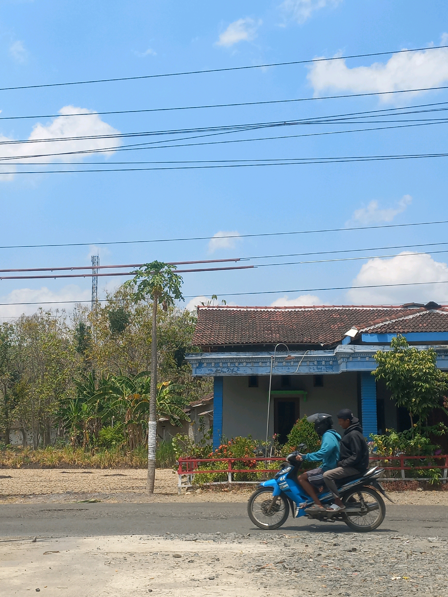 Two individuals, one wearing a blue hoodie and the other a black hoodie and cap, ride a blue motorcycle. The motorcycle is traveling on a paved road, which transitions to a gravel shoulder. In the background, there is a building with a tiled roof and blue trim, situated behind a red and white fence. A tall, thin tree stands to the left of the building, and beyond that are dense trees and foliage. A cellular tower is visible in the distance. The sky above is clear blue with scattered white clouds. The scene appears to be in a semi-rural or suburban area.