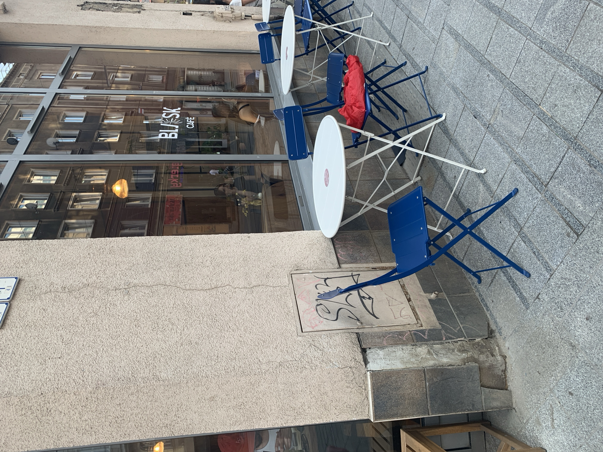 The image depicts an outdoor seating area of a cafe, identified as "BLASK CAFE", located in Poznan, Poland. The scene is set on a paved sidewalk in front of a building with large glass windows reflecting the urban environment. Several round white tables with white foldable metal frames are arranged on the sidewalk. Blue foldable metal chairs are placed around these tables. One chair has a crumpled red bag on it. The building facade is textured, and a gray electrical box with graffiti is visible on the wall near the tables. The ground is paved with gray square tiles. The reflection in the windows shows an urban street with buildings and people. The overall atmosphere suggests a casual outdoor dining or coffee area.